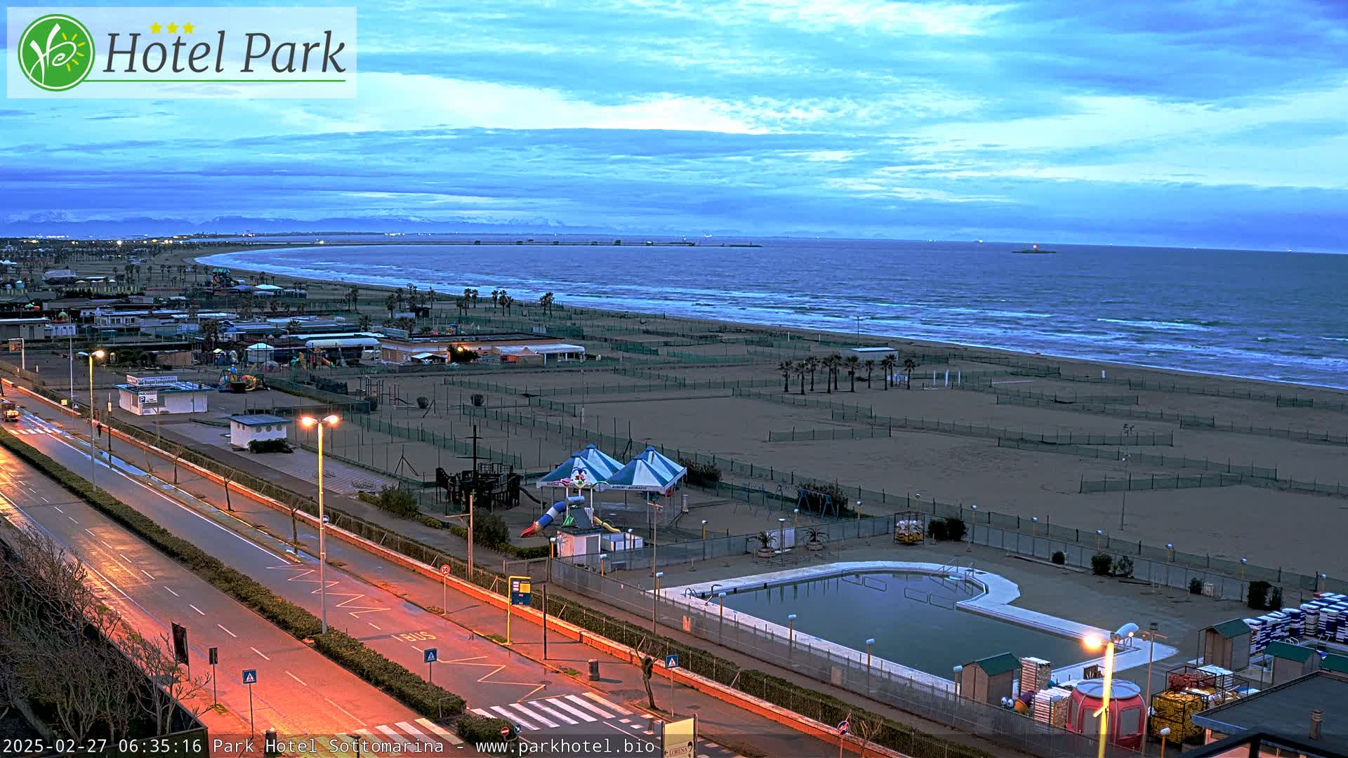 An empty beach with a playground, swimming pool, and road next to it is visible at dawn under a mostly clear, twilight sky.