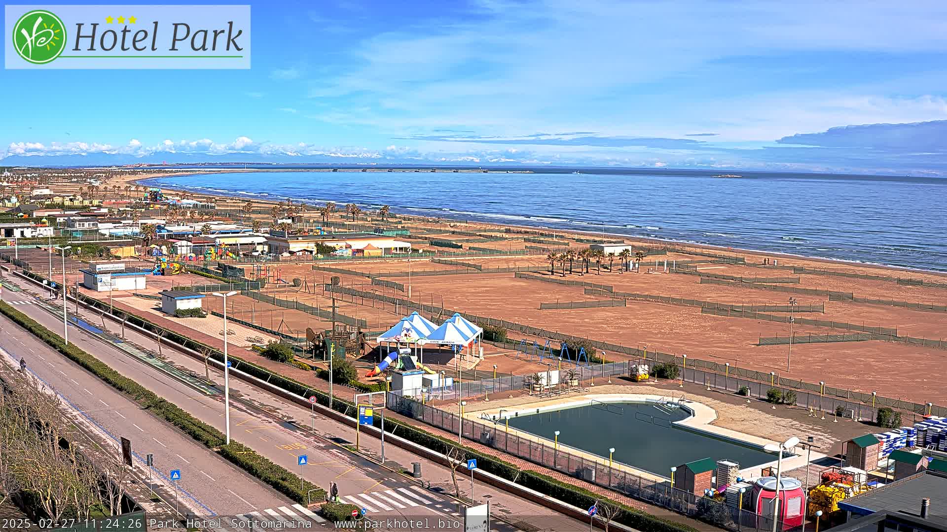 A beachside resort area with a swimming pool, playground equipment, and tennis courts is visible under a mostly clear sky, overlooking a wide sandy beach and calm ocean.