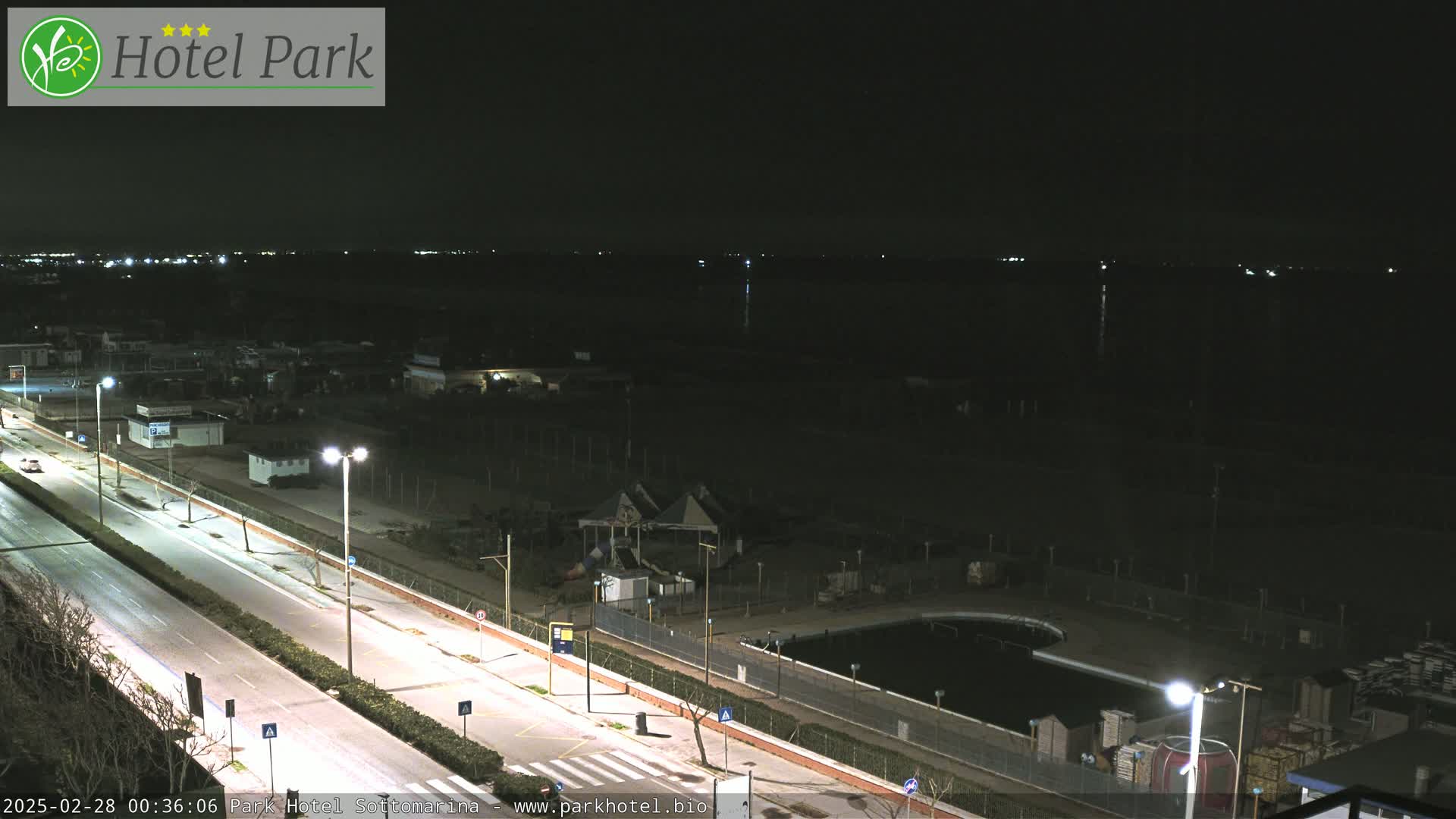 A nighttime view of a mostly empty roadway and beachfront area with a dark body of water in the background, under clear skies.