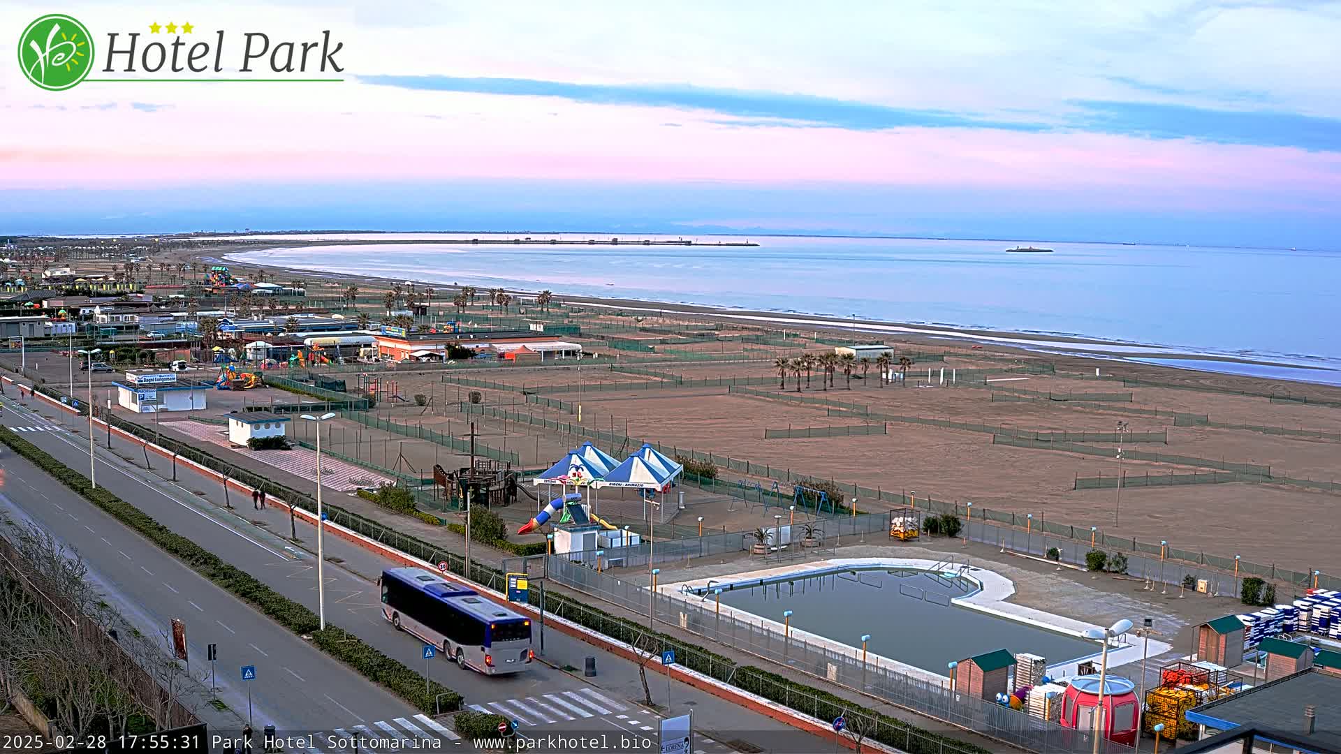 A wide view of a beachside resort area under a pale pink and blue twilight sky shows a sandy beach, a swimming pool, playground equipment, a road with a bus, and calm ocean waters in the distance.