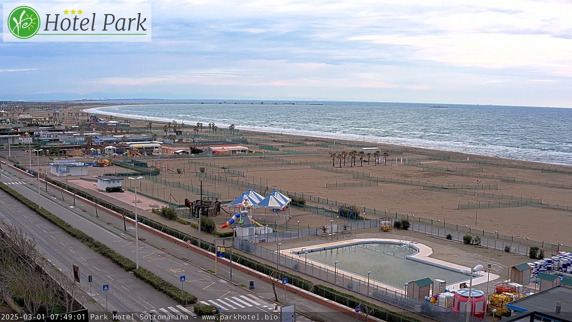 An outdoor view shows a sandy beach with a calm sea, a road next to a playground, and a swimming pool adjacent to the beach area on an overcast day.