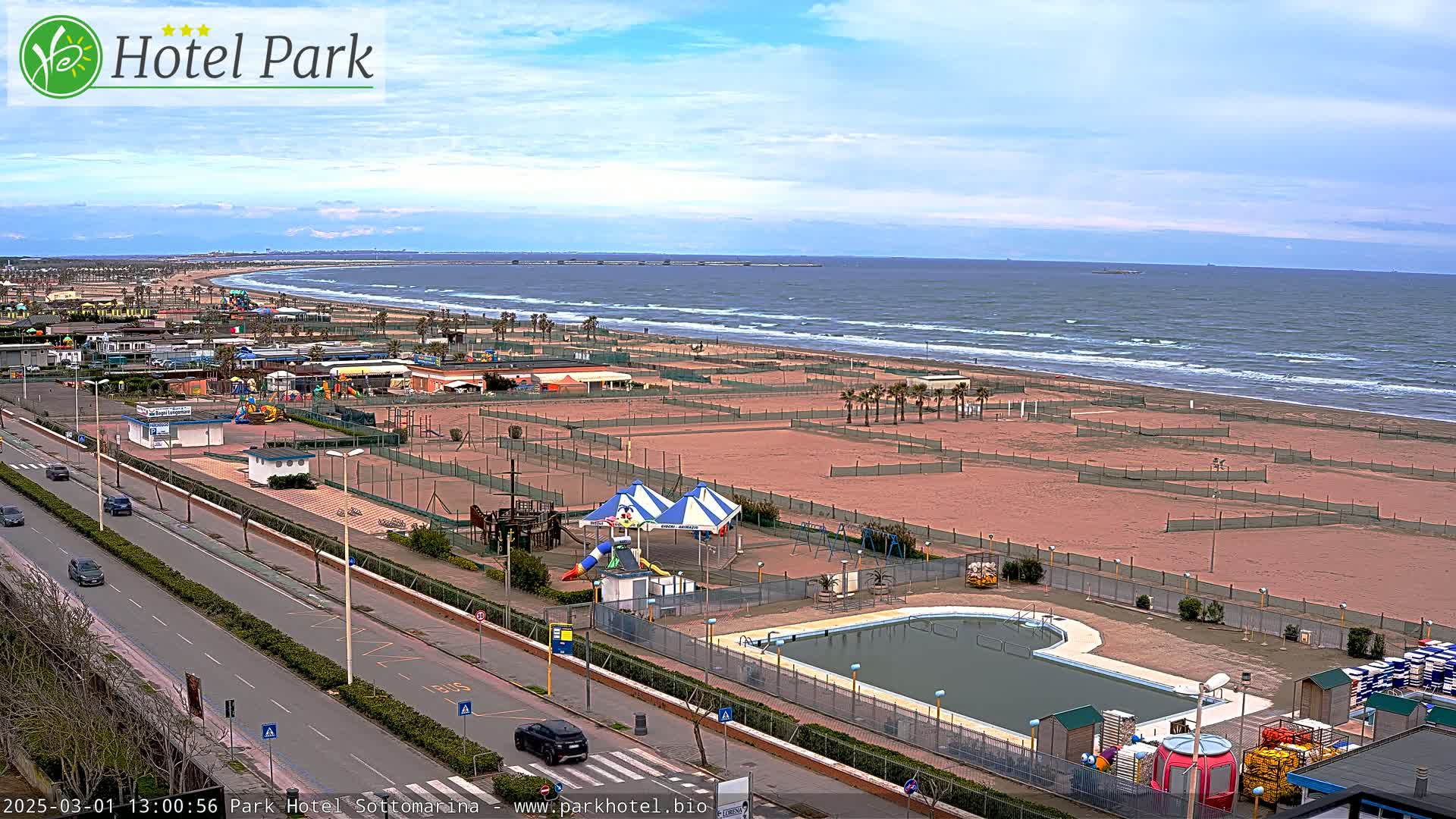 A beachside amusement park with a pool is visible, overlooking a wide sandy beach and a somewhat choppy sea under a mostly cloudy sky.