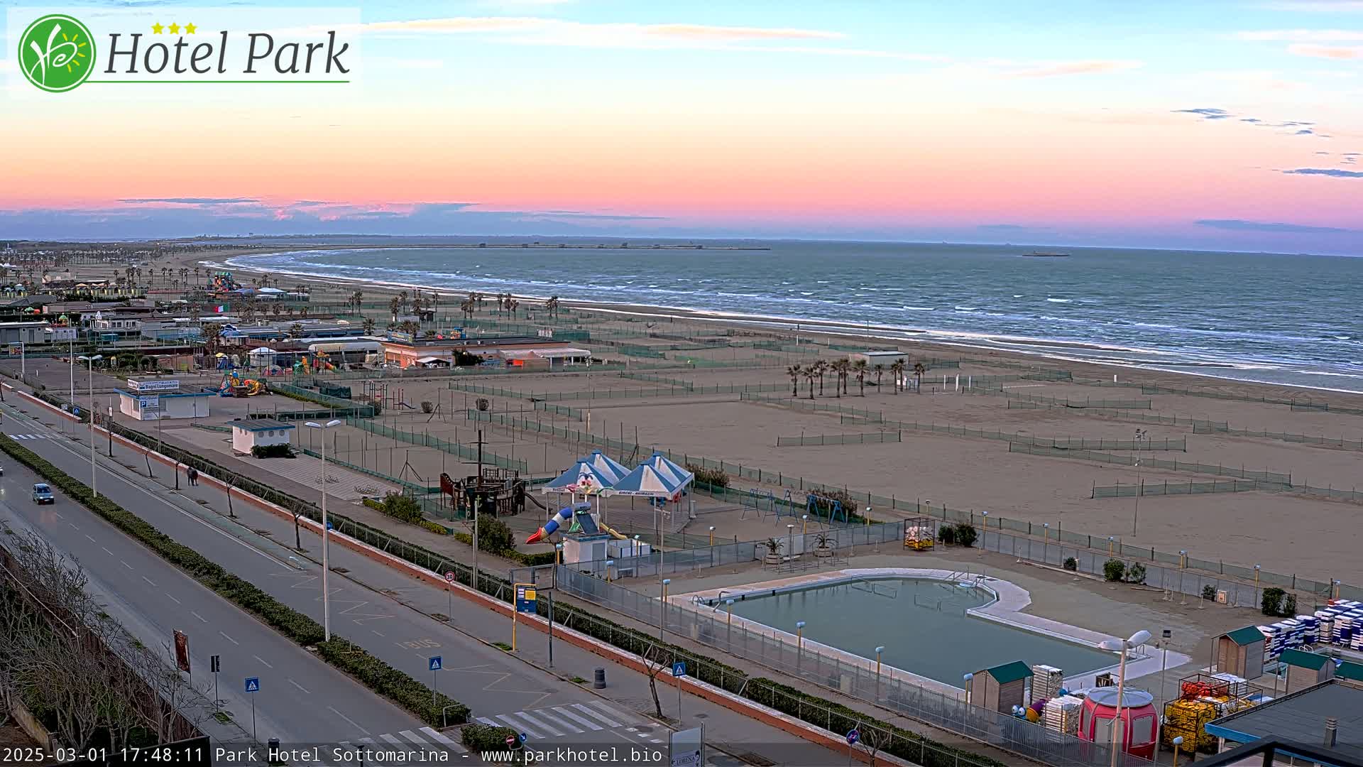 An aerial view shows a beach with a calm sea under a pink and orange sunset, featuring a beachfront area with a swimming pool, playground equipment, and a roadway.