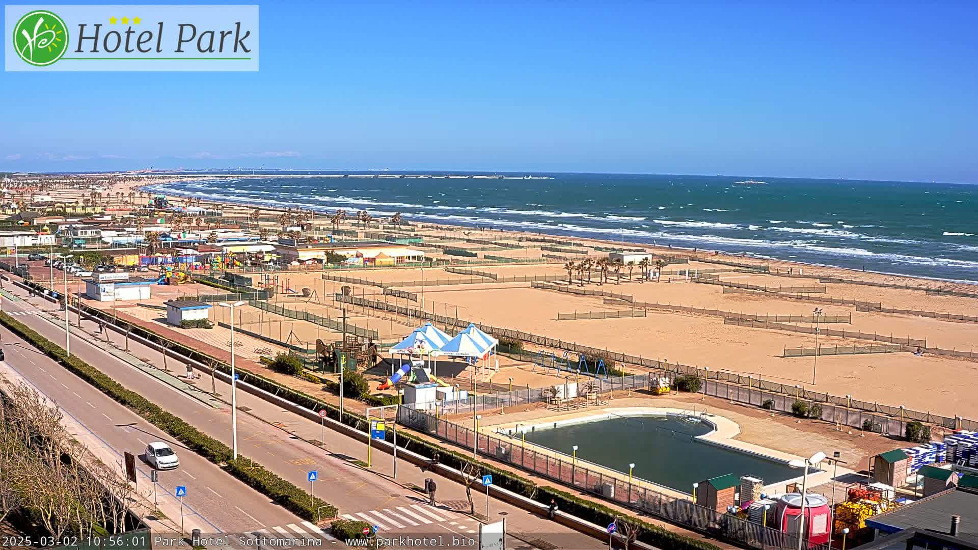 A sandy beach with a playground, swimming pool, and volleyball courts is visible, alongside a road and ocean with moderate waves under a clear blue sky.