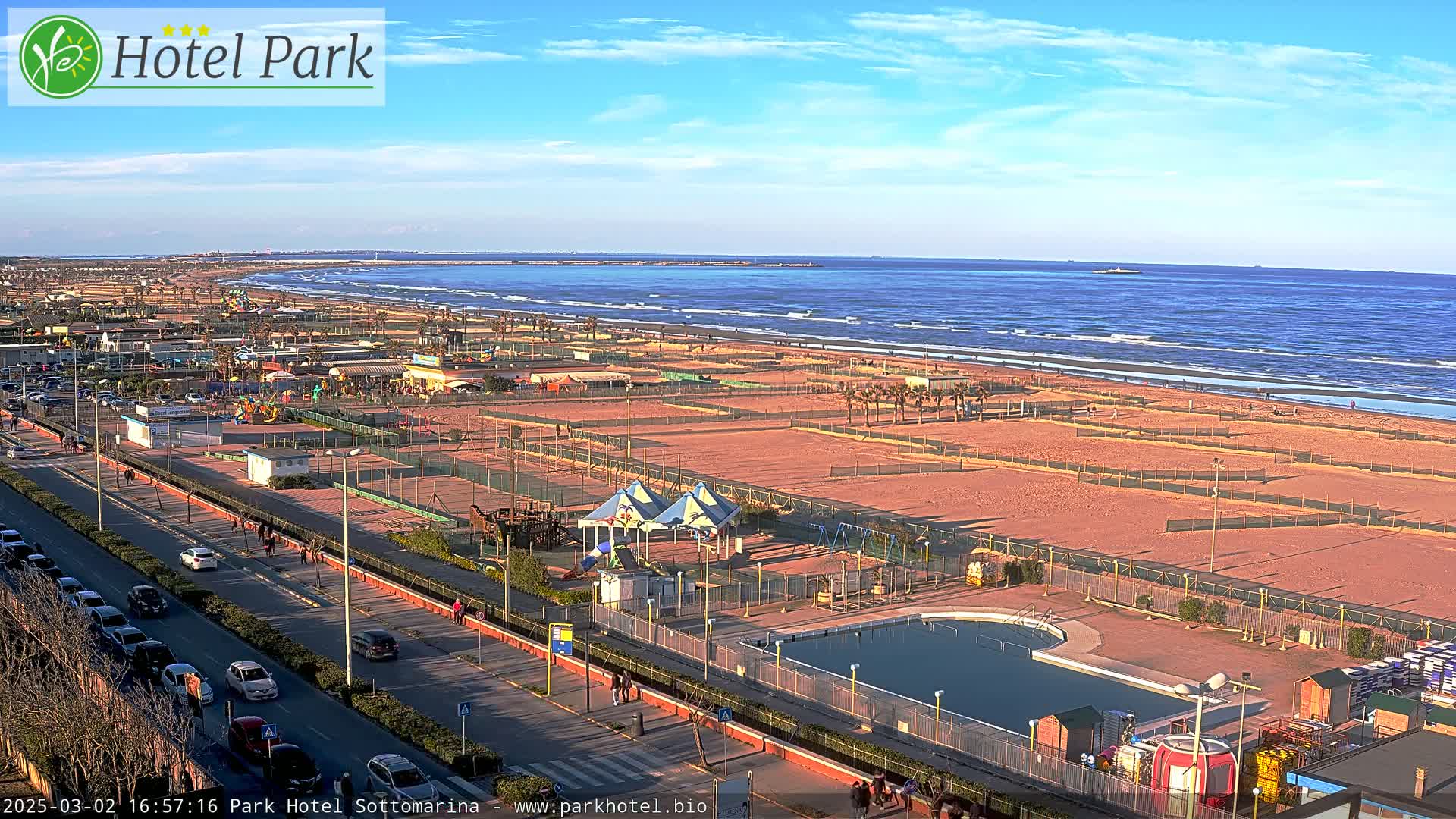 An aerial view shows a beach with volleyball courts, a swimming pool, playground equipment, and a roadway with parked cars, all under a mostly clear, sunny sky.
