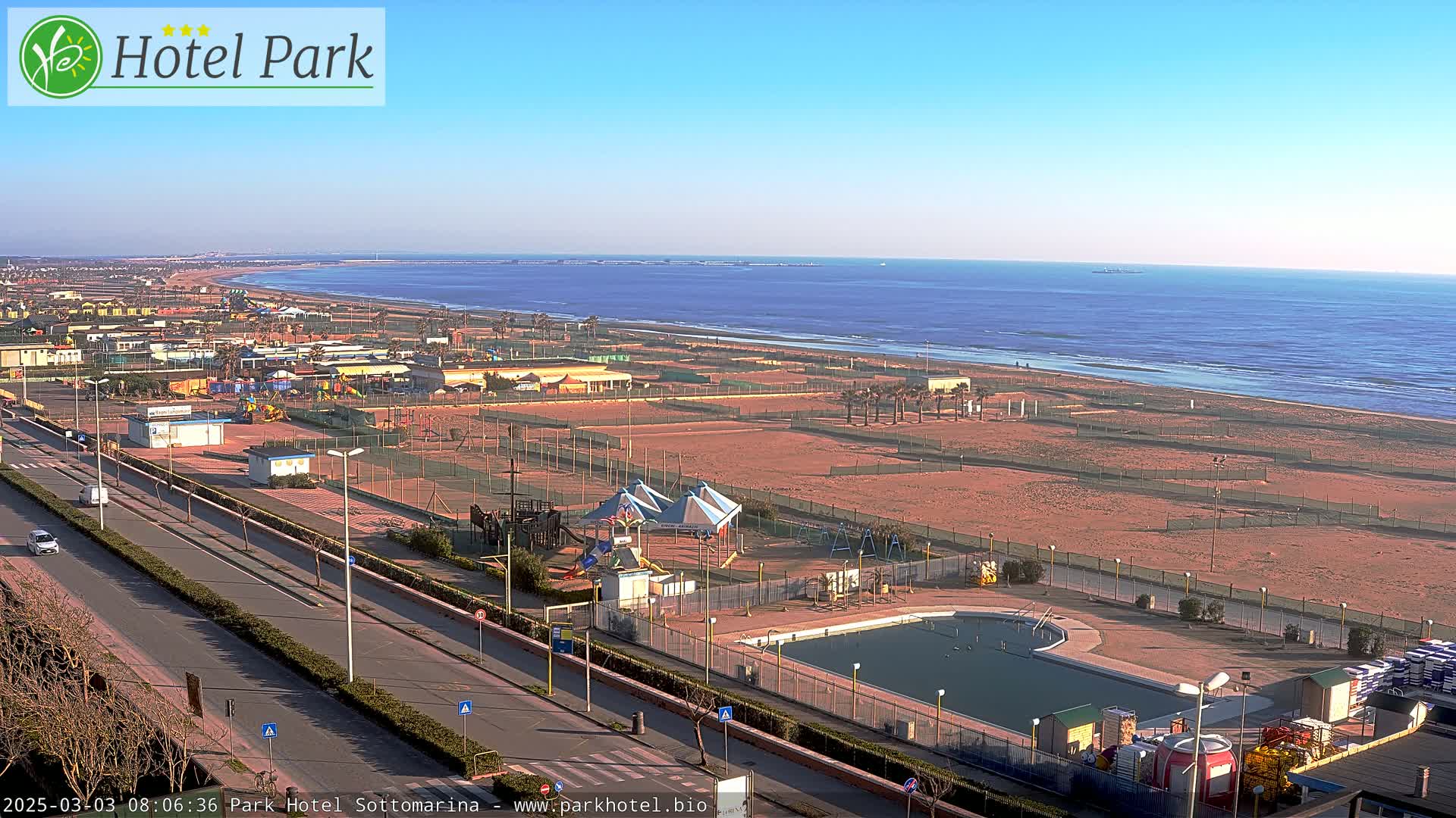A high-angle, long shot shows a beachside resort area on a sunny day with a calm ocean in the background;  a swimming pool and playground are visible in the foreground near a road.