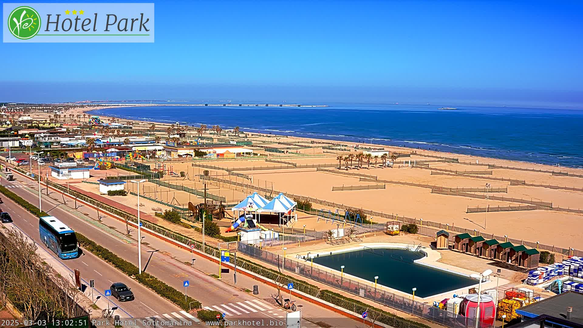 A beachside resort on a sunny day, featuring a swimming pool, playground, and volleyball courts, with a bus and cars on a road nearby.