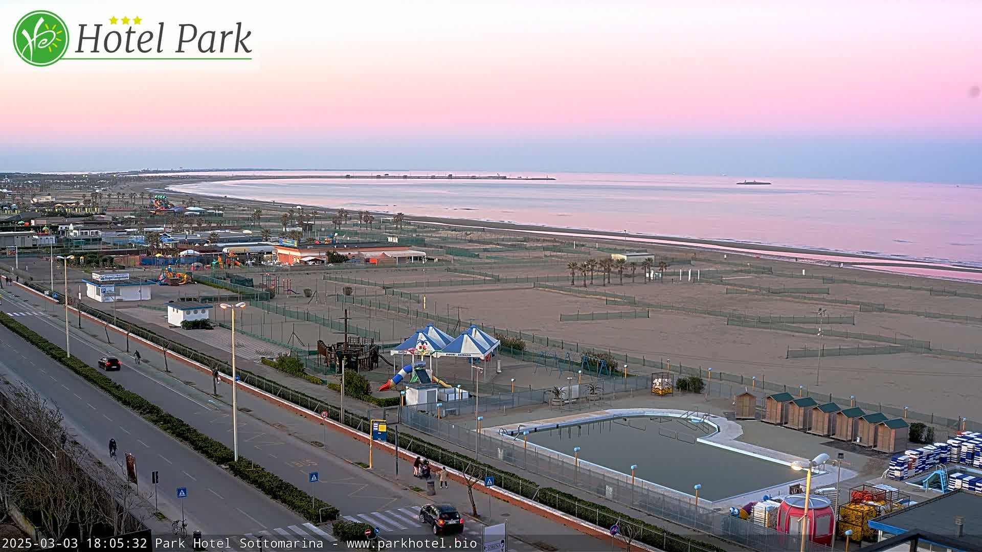 A beachside amusement park with a swimming pool is seen at dusk under a pink and purple sky.