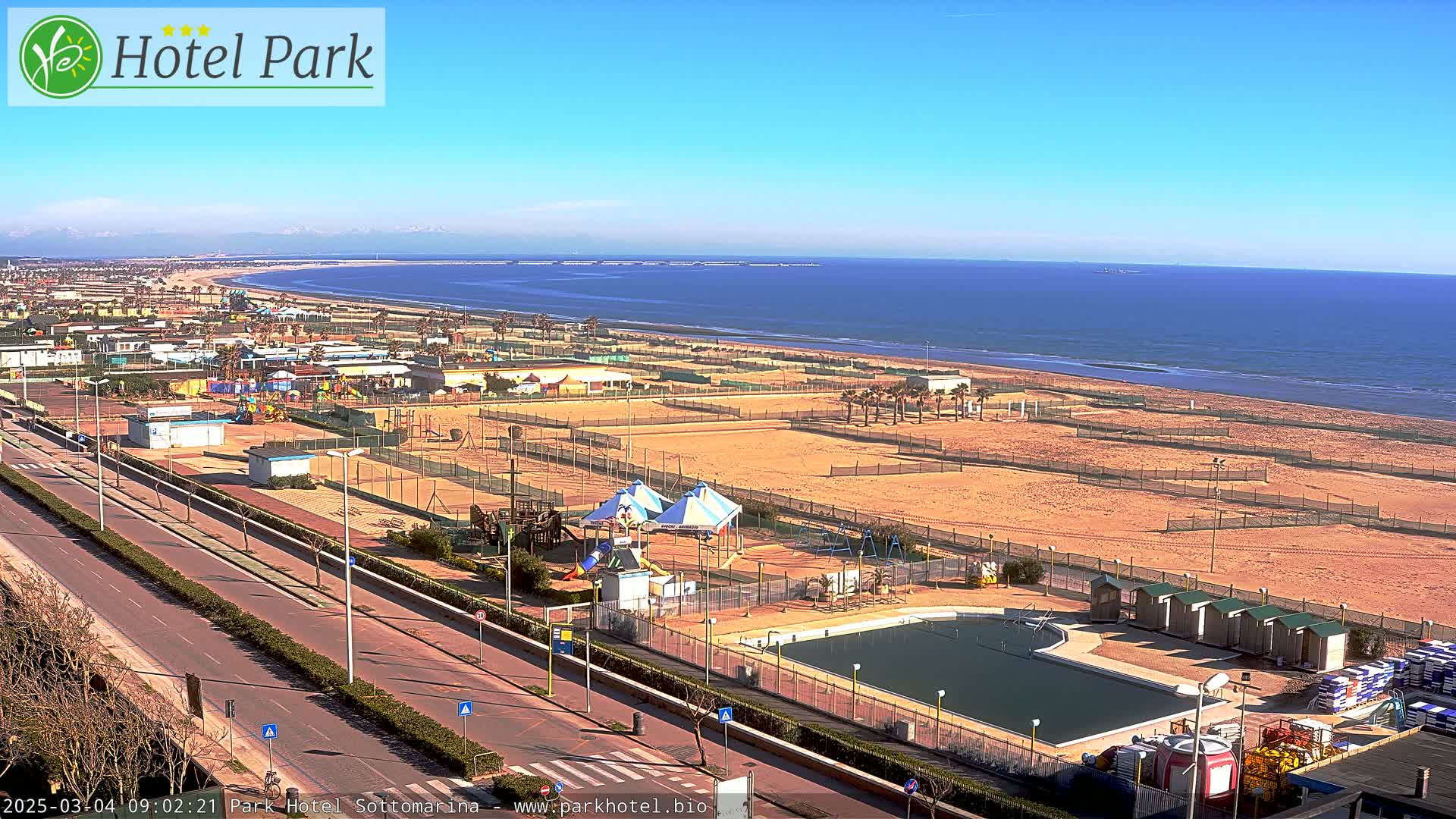 A beachside area with a pool, playgrounds, and volleyball courts is visible under sunny skies, with a calm ocean in the background.