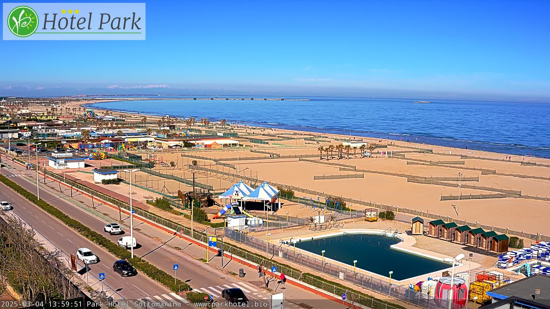 A beachfront resort area with a swimming pool, playground, and volleyball courts under a clear blue sky.