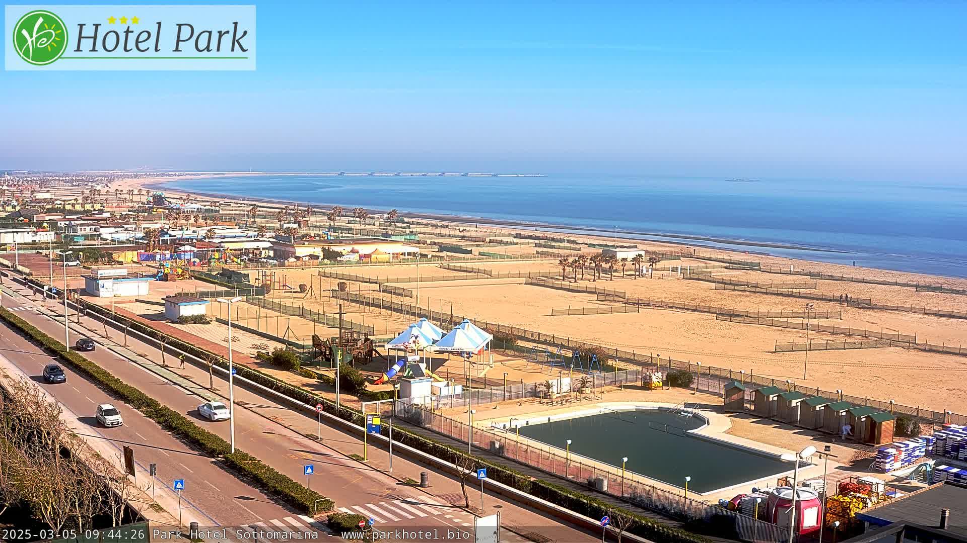 An aerial view shows a beachside resort with a swimming pool, playground, and volleyball courts on a sunny day, overlooking a calm ocean.