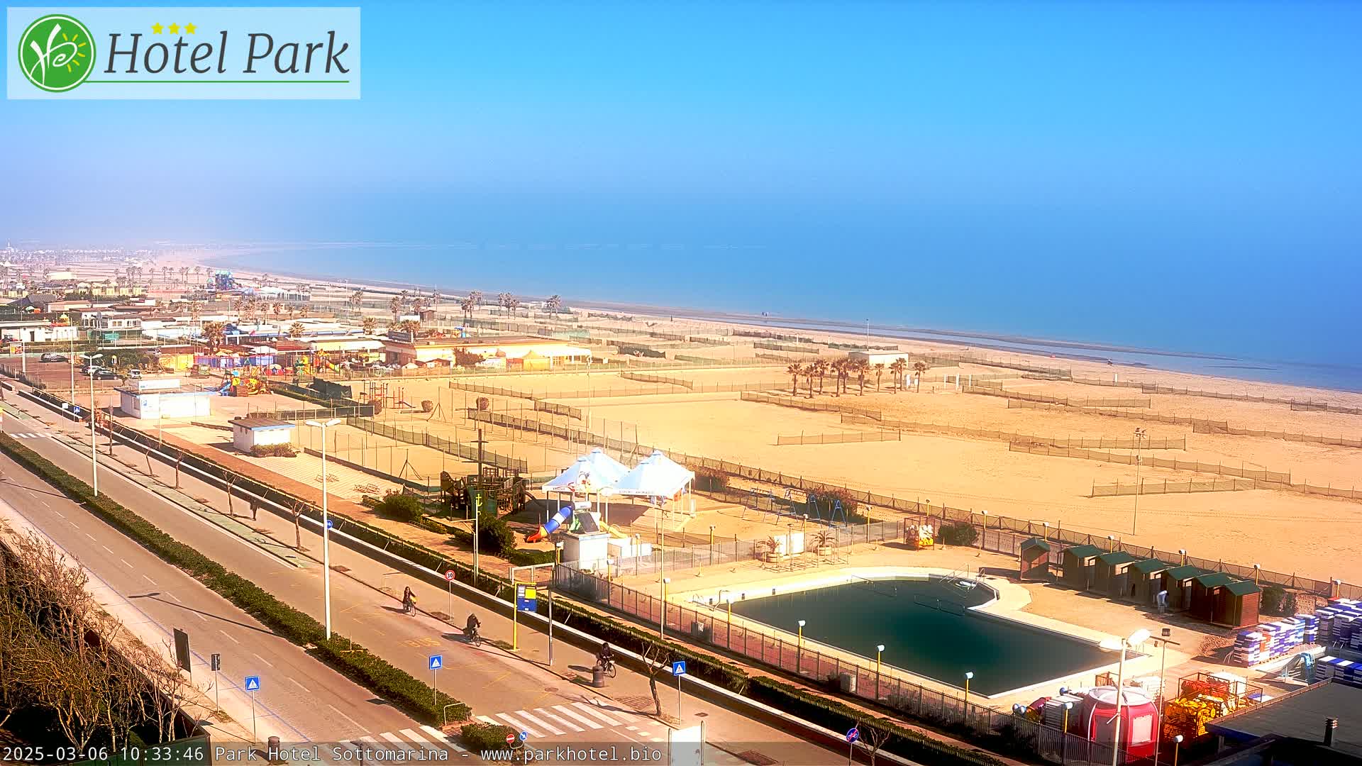 A sunny day overlooking a beach with a swimming pool, playground, and tennis courts next to a road.