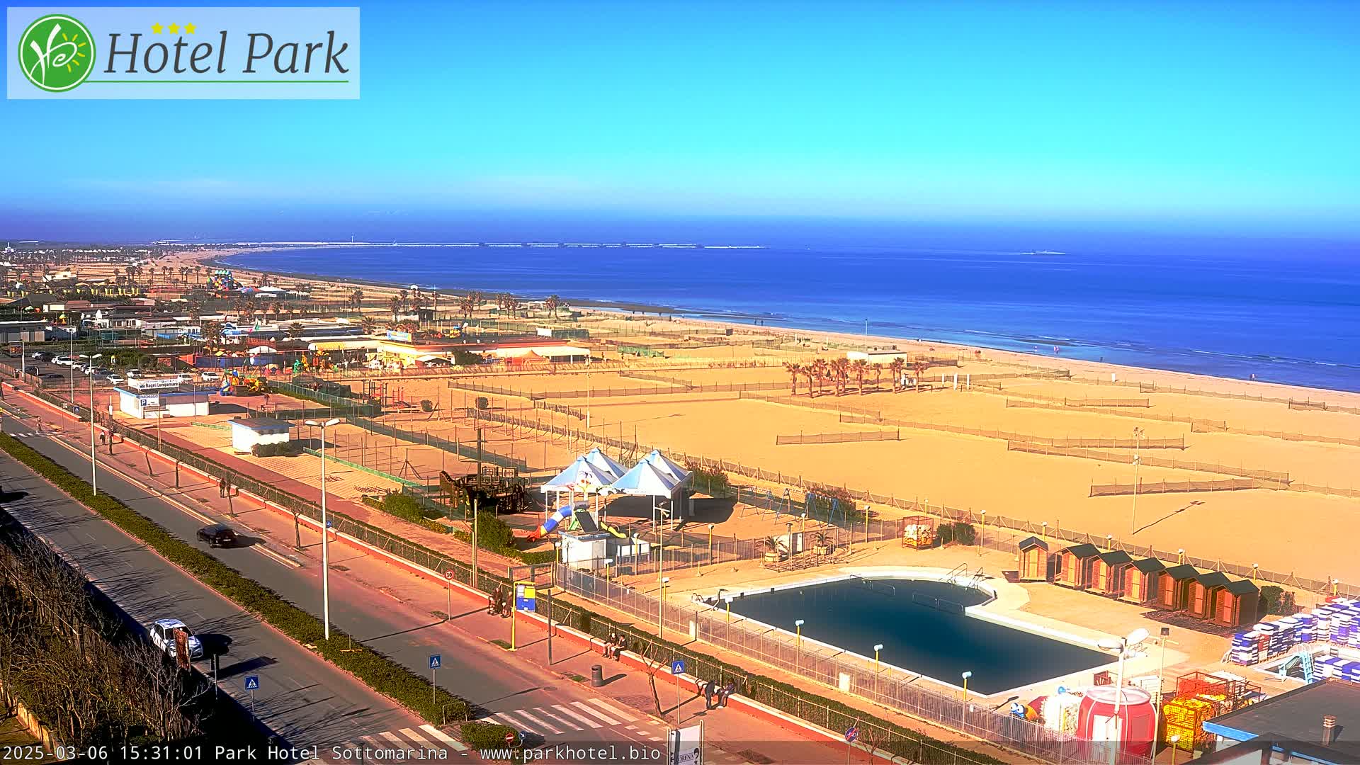 A sunny beach scene shows a sandy shoreline with volleyball courts, a swimming pool, and a playground, all situated next to a road and overlooking a calm ocean.