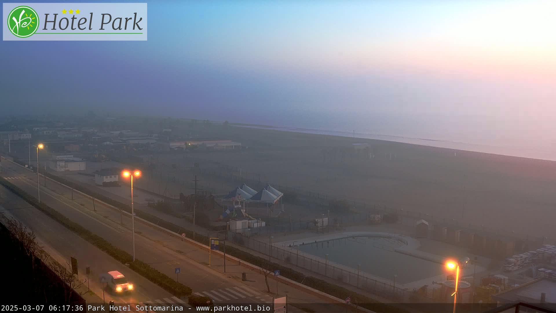A hazy sunrise illuminates a beach, a road with cars, and a resort area with a swimming pool and playground.
