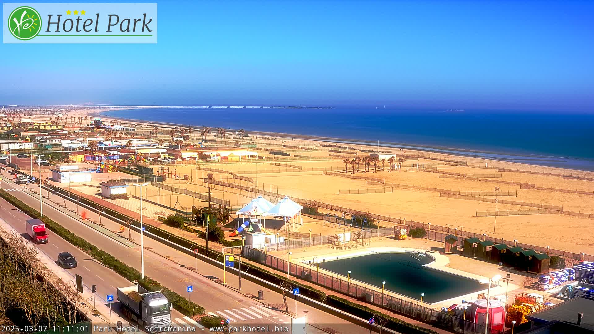 A beachside resort area on a sunny day, featuring a swimming pool, playground equipment, and volleyball courts, with a calm ocean in the background.