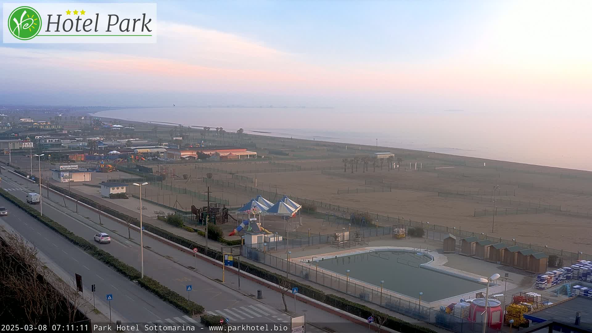 A hazy sunrise illuminates a seaside resort with a swimming pool, playground, and sandy beach.