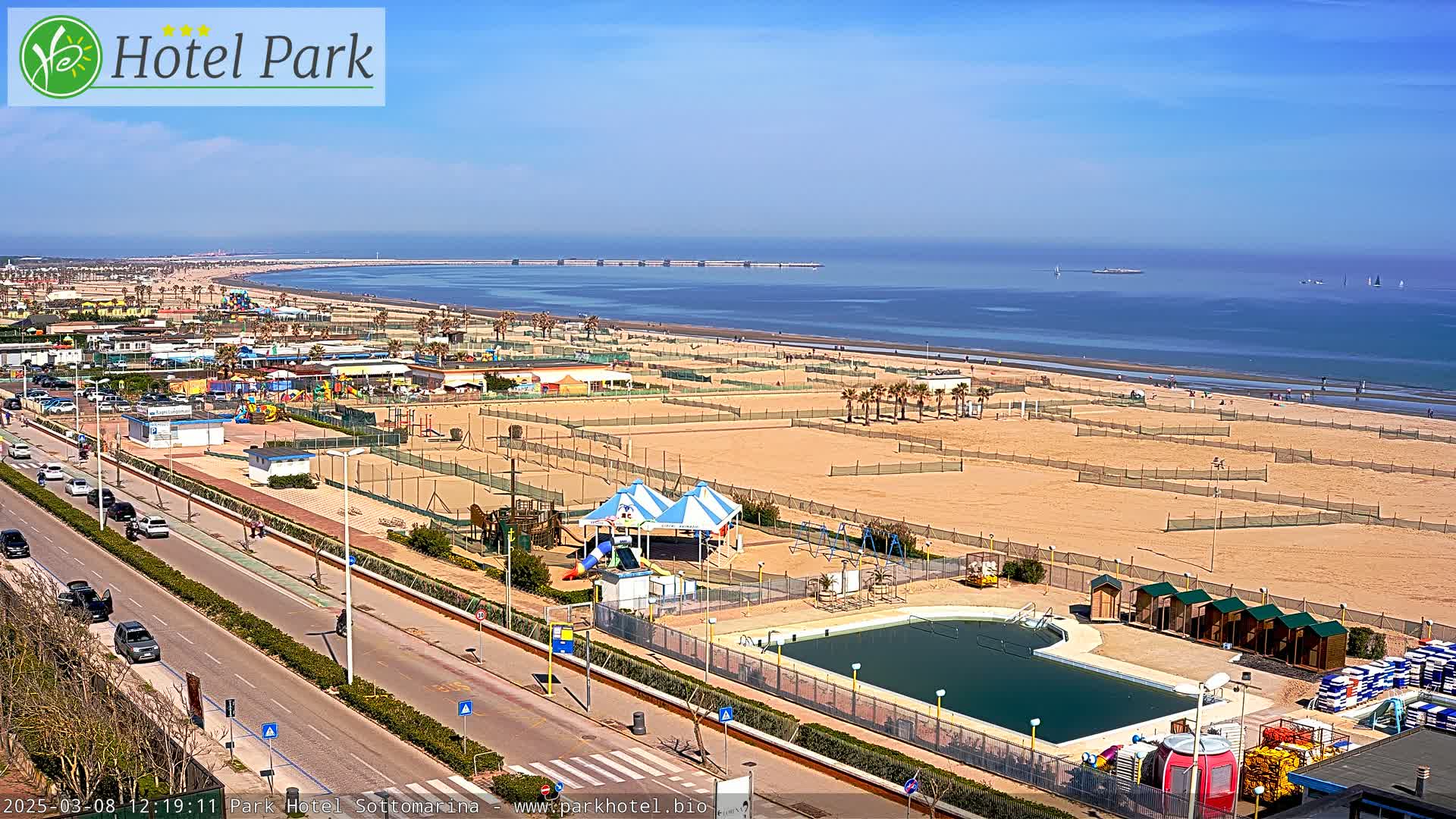 A sunny day at a beach resort shows a sandy beach with volleyball courts, a swimming pool, playground equipment, and a road with cars.