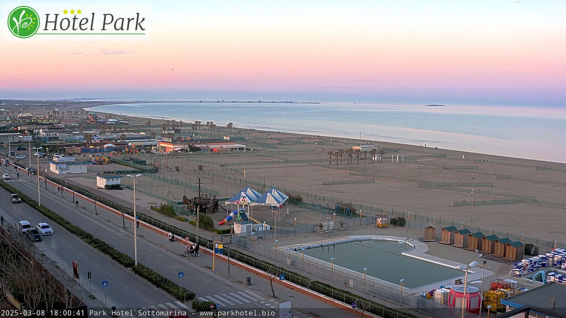 A beachside view at sunset shows a calm ocean, a sandy beach with playground equipment and a swimming pool, and a road with cars.
