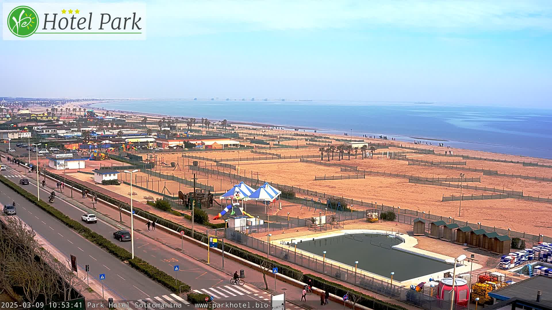 A sandy beach with a playground, swimming pool, and a road with cars is visible, overlooking a calm sea under a clear sky.