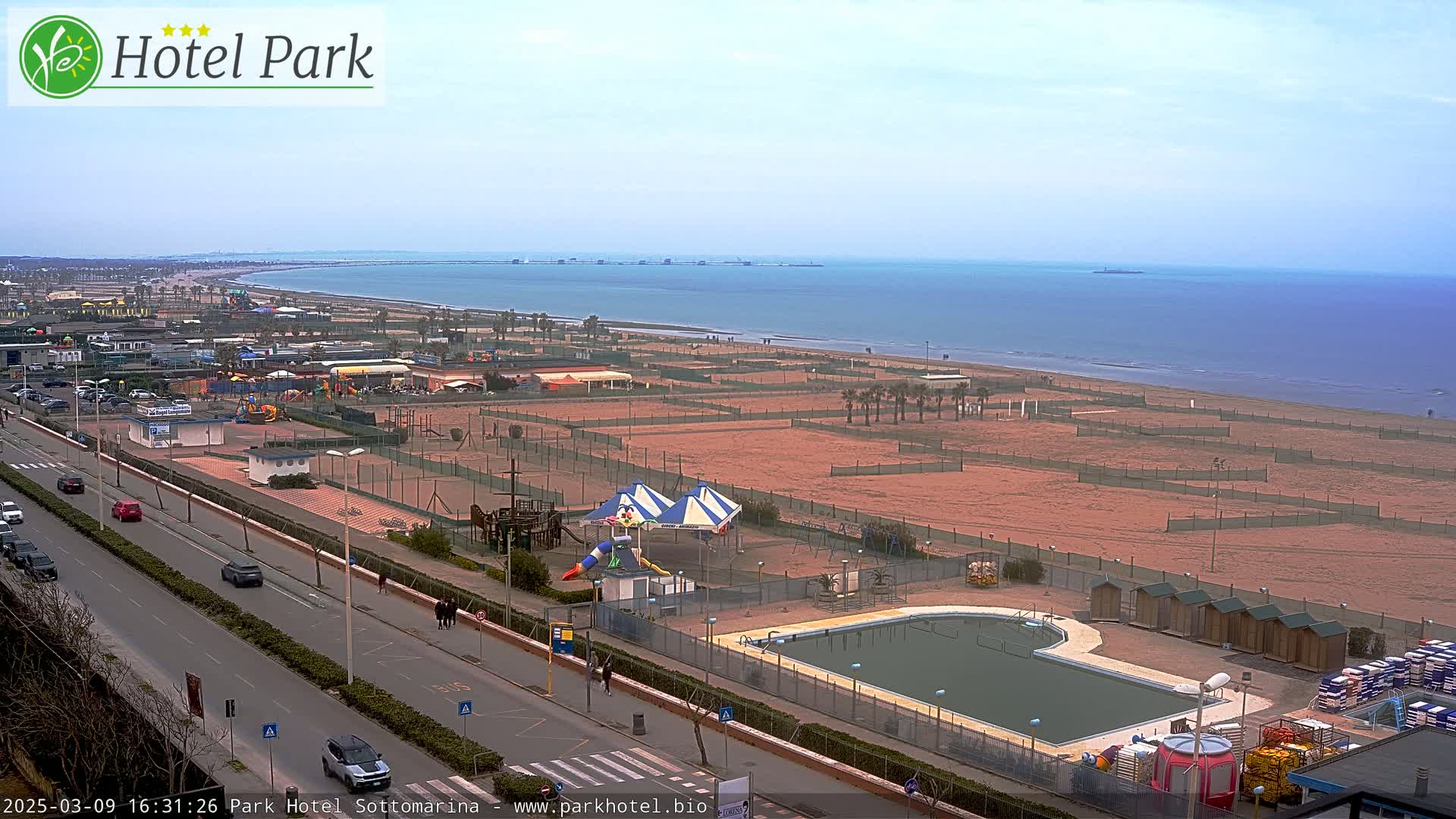 An aerial view of a beachside resort area on an overcast day, showing a sandy beach, a swimming pool, playground equipment, and a road with cars.