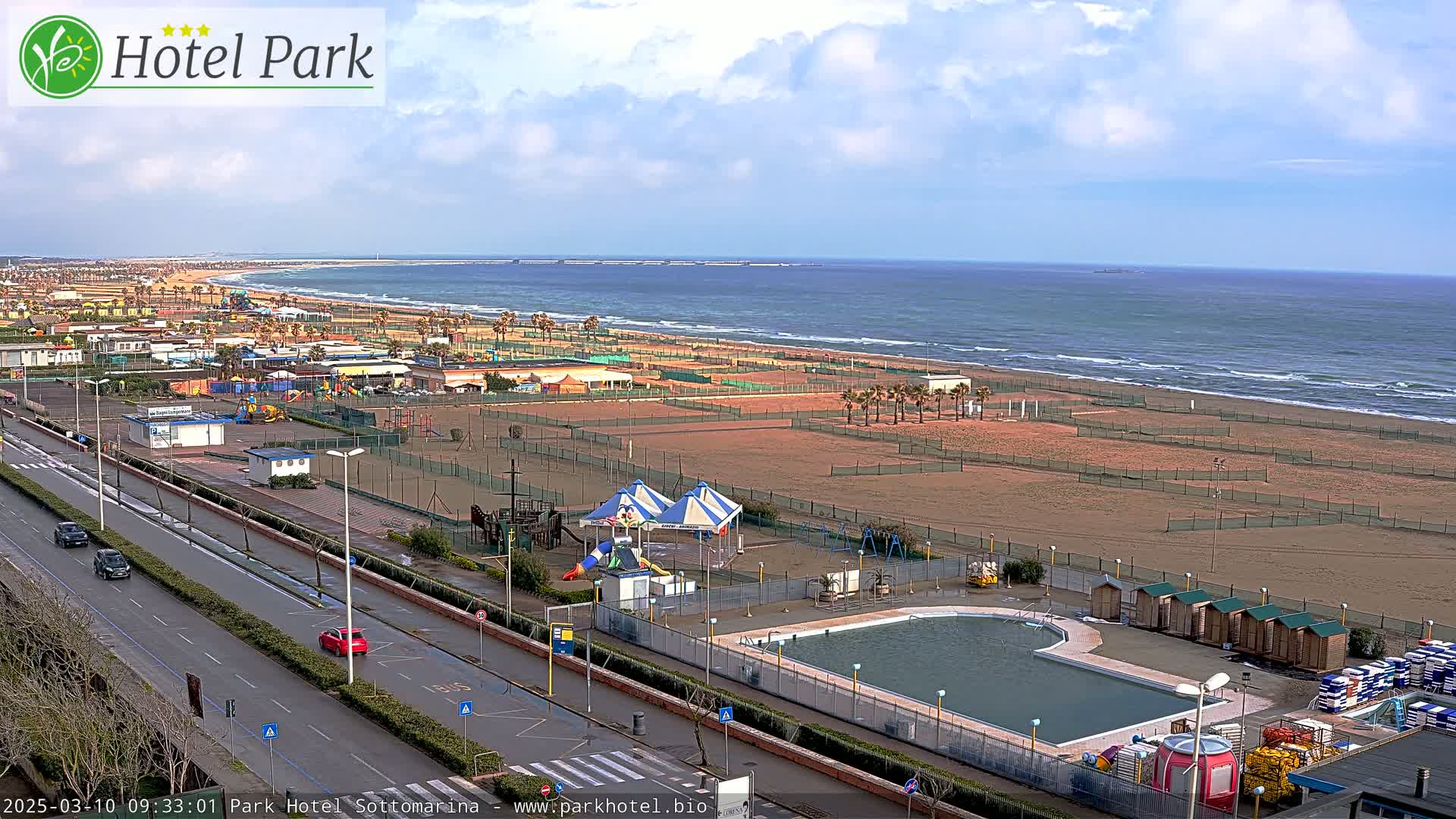 A sandy beach with a calm ocean, a swimming pool, playground equipment, and a road with cars are visible under a partly cloudy sky.