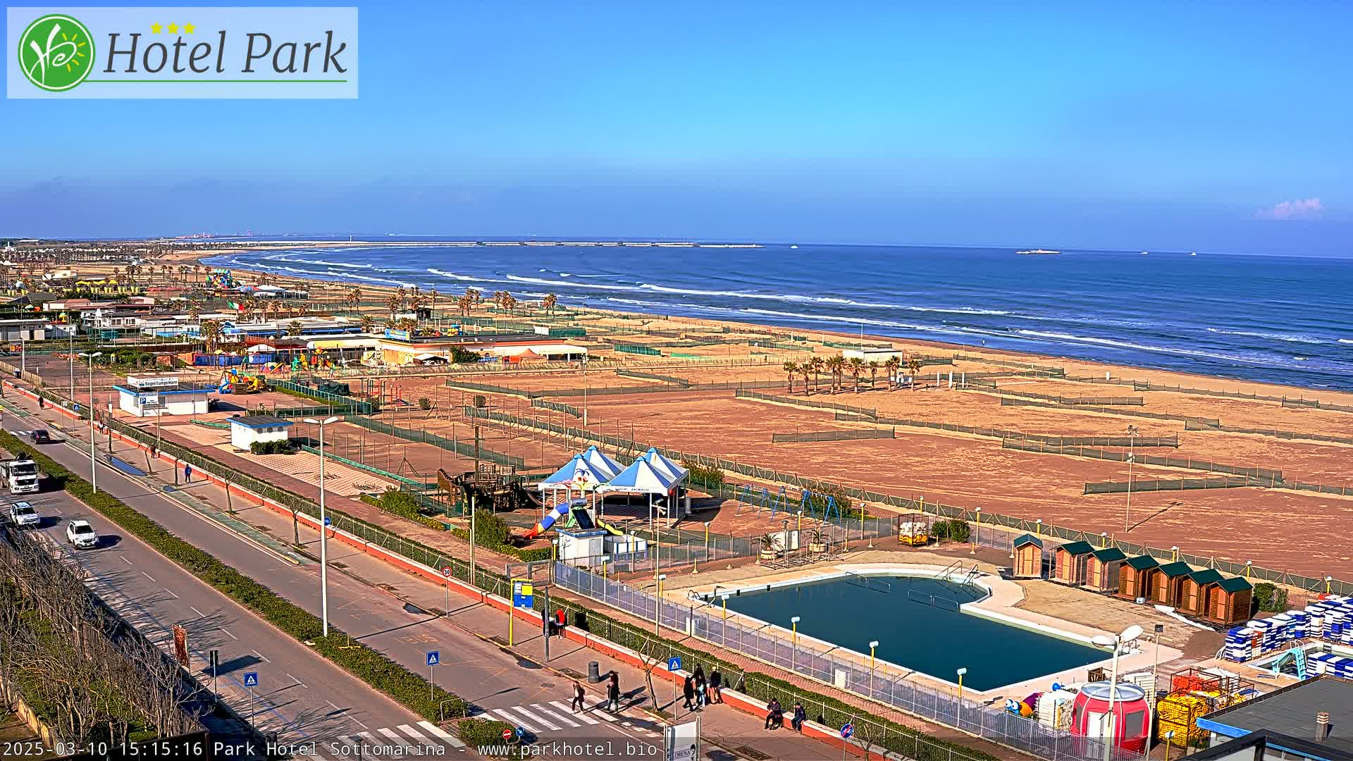 A beachside resort area with a swimming pool, playground equipment, and beach volleyball courts under a clear, sunny sky.