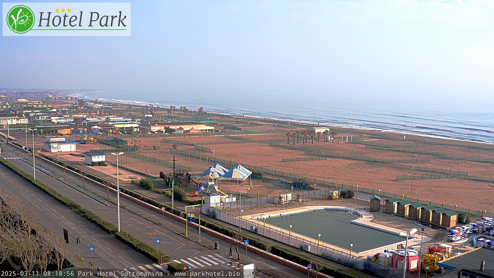 A beachside area with a swimming pool, playground, and sandy beach under a clear, hazy sky.