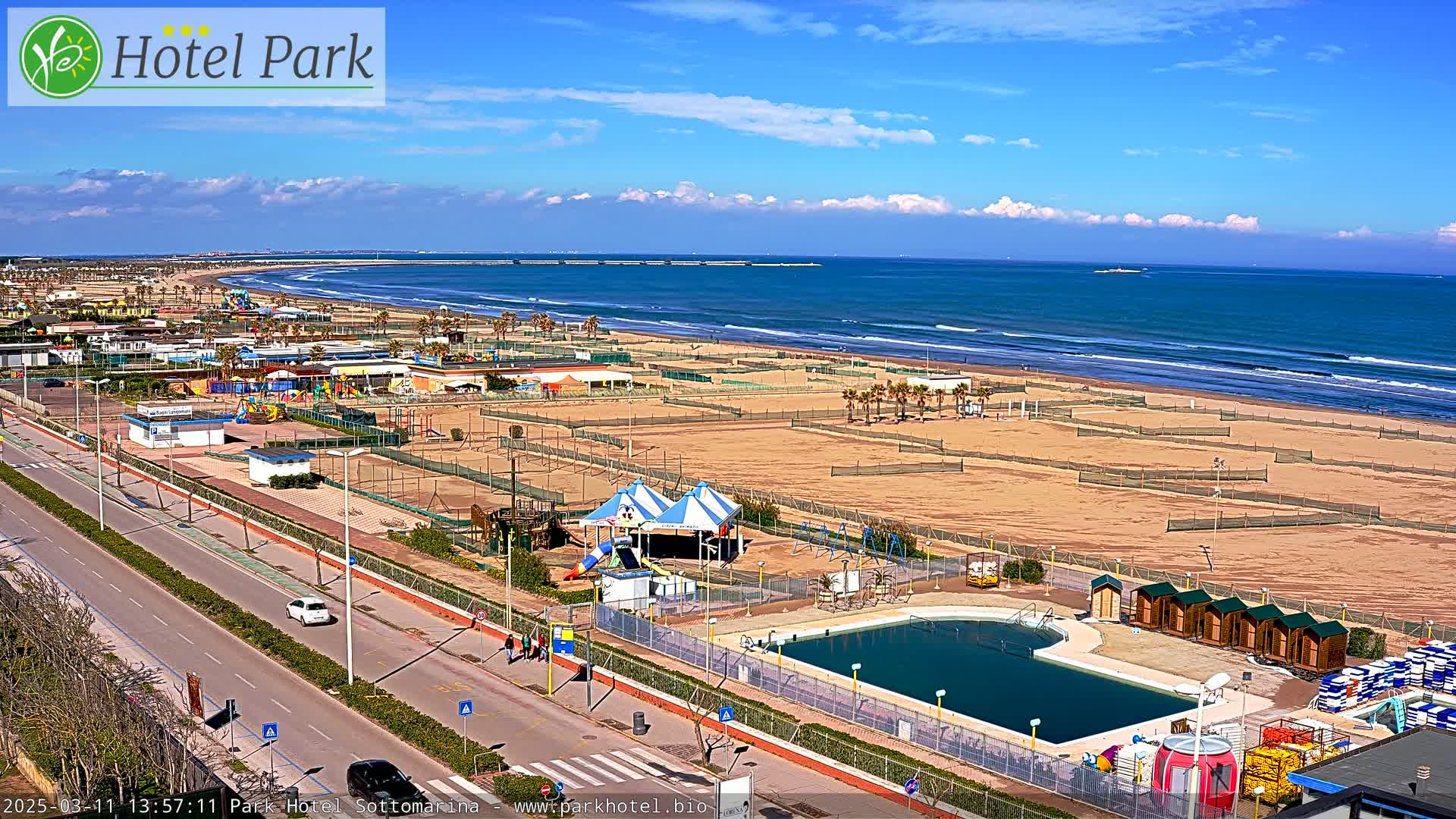 A beachfront area with a swimming pool, playground equipment, and volleyball courts under a sunny sky with some clouds.