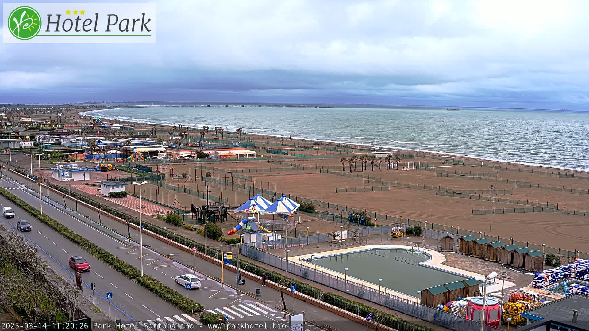 An overcast day reveals a beach with a swimming pool, playground equipment, and a road with cars, all situated next to a calm sea.