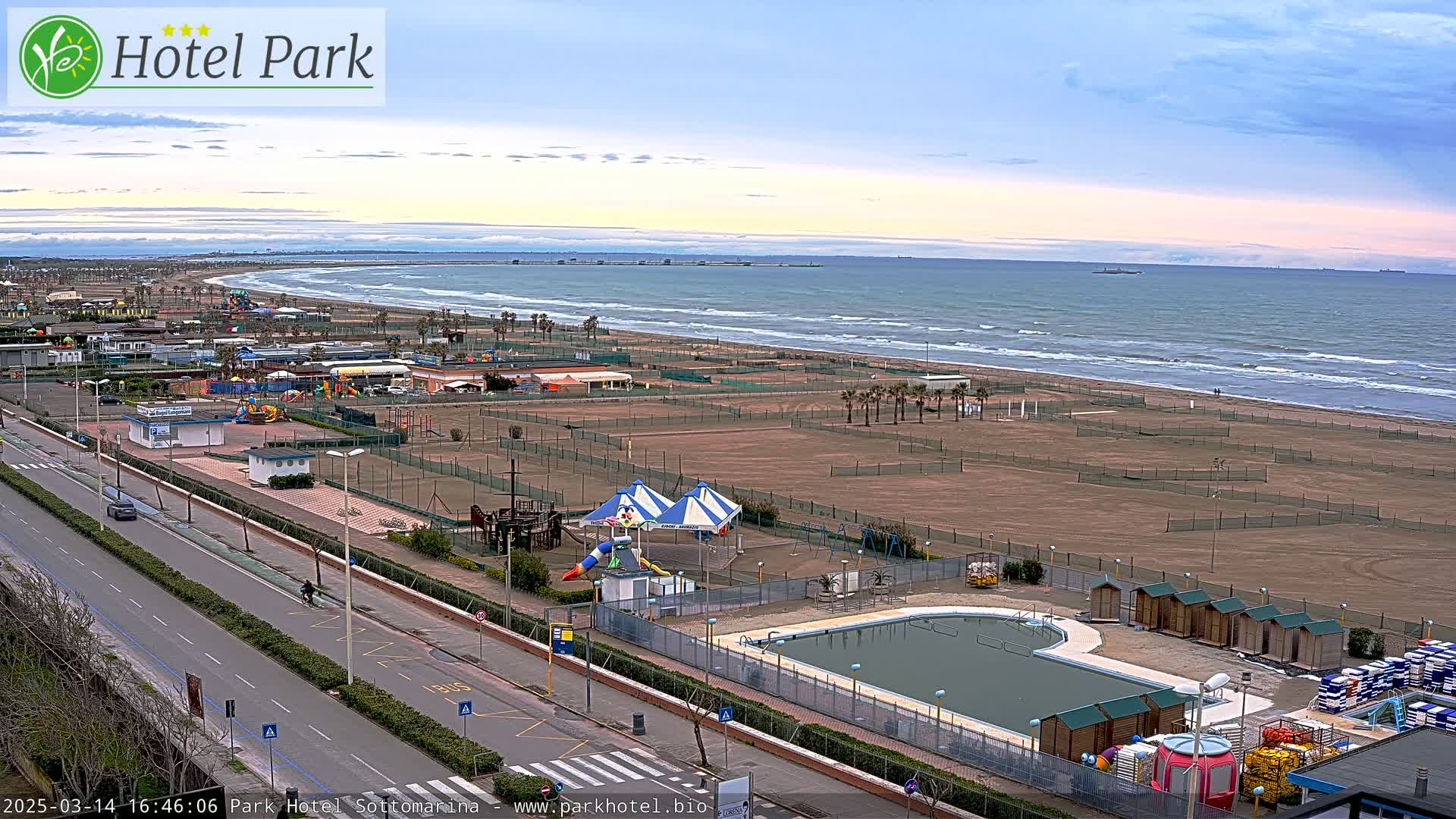 An aerial view shows a beach with a calm sea under a partly cloudy sky, featuring a resort area with a pool, playground, and tennis courts.