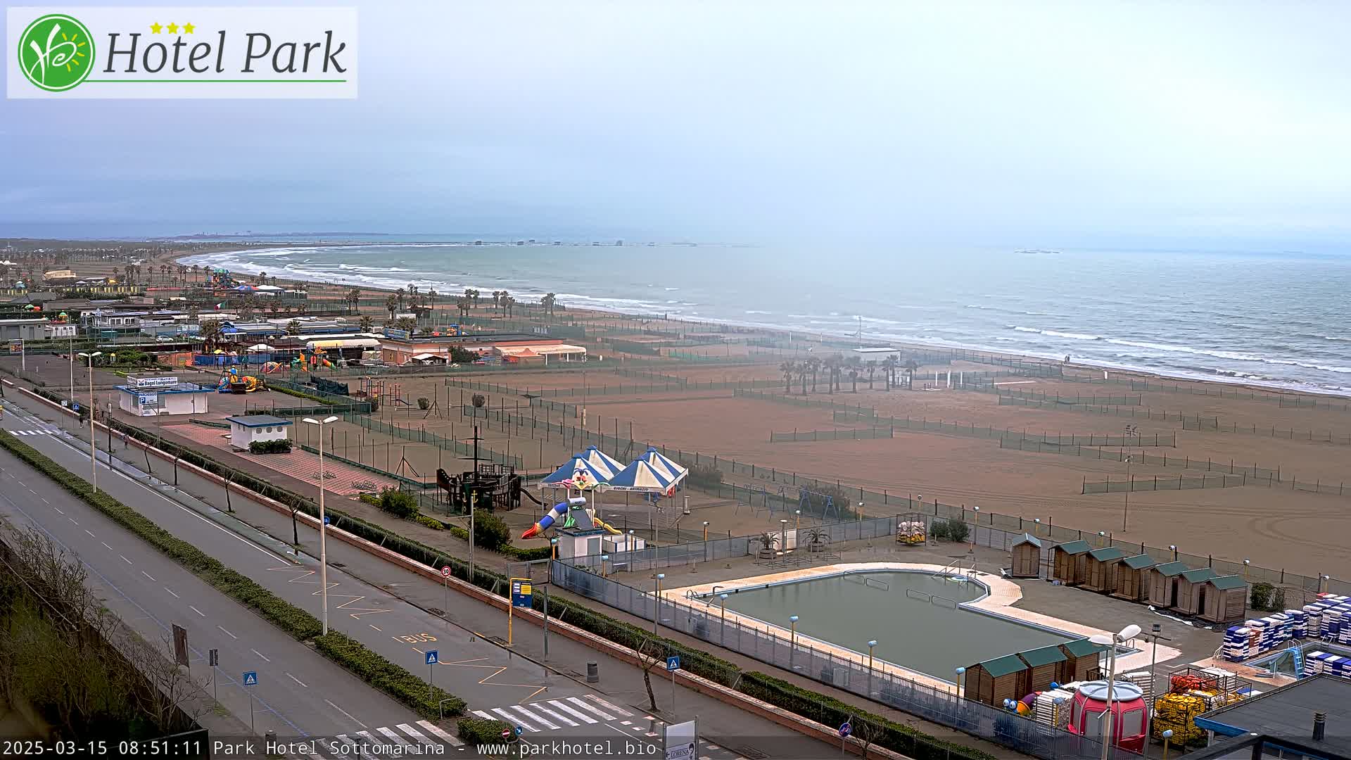 An overcast day reveals a beachside resort with a pool, playground equipment, and volleyball courts, all situated next to a wide sandy beach with gently rolling waves.
