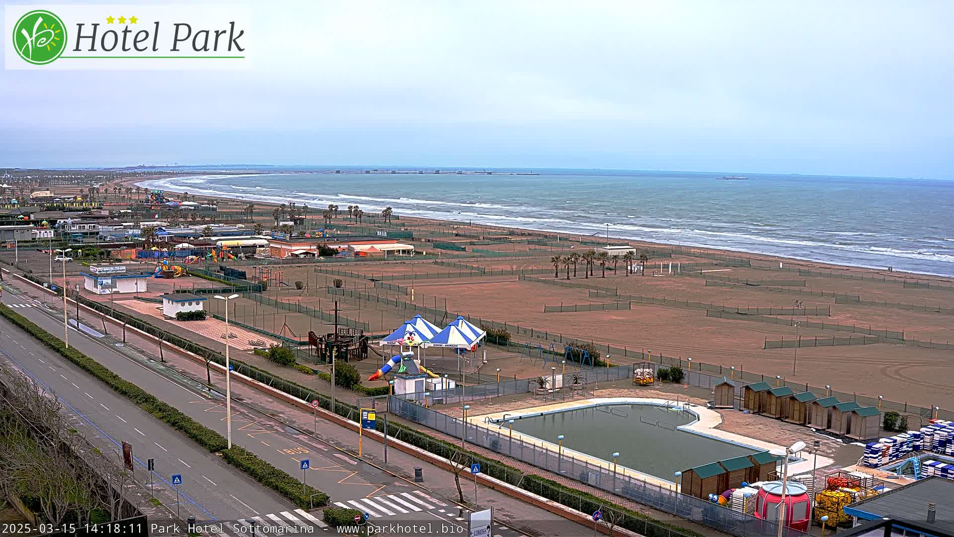 A wide view of a beach with a pool, playground, and other recreational facilities, under an overcast sky.