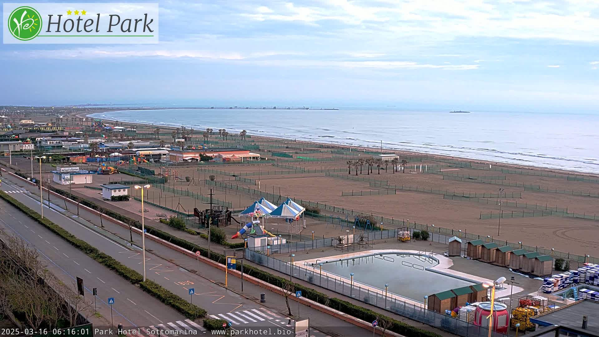 An aerial view shows a beach with a swimming pool, playground, and sandy expanse under a partly cloudy sky.