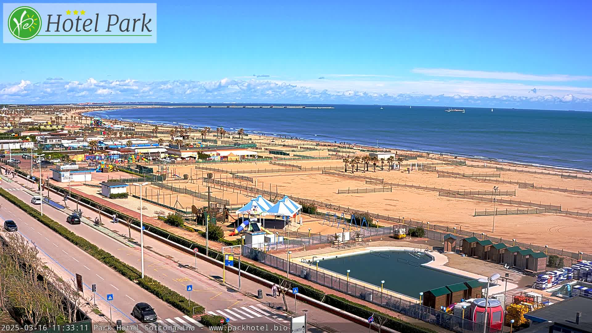 A beach with volleyball courts, a swimming pool, playground equipment, and a road is seen from an elevated viewpoint on a sunny day with some clouds.