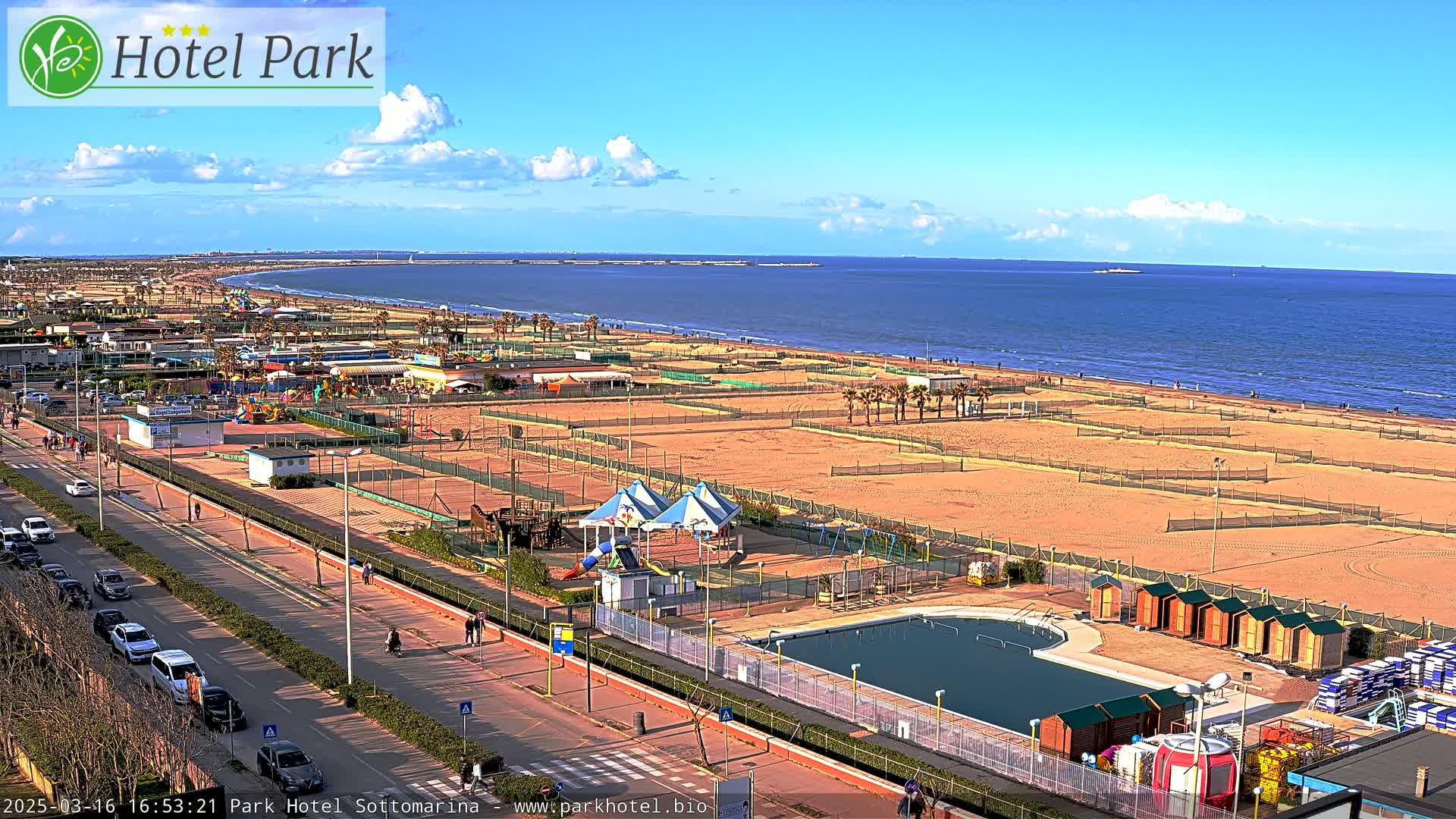A sunny day overlooking a beach with volleyball courts, a swimming pool, playground equipment, and a road lined with cars.