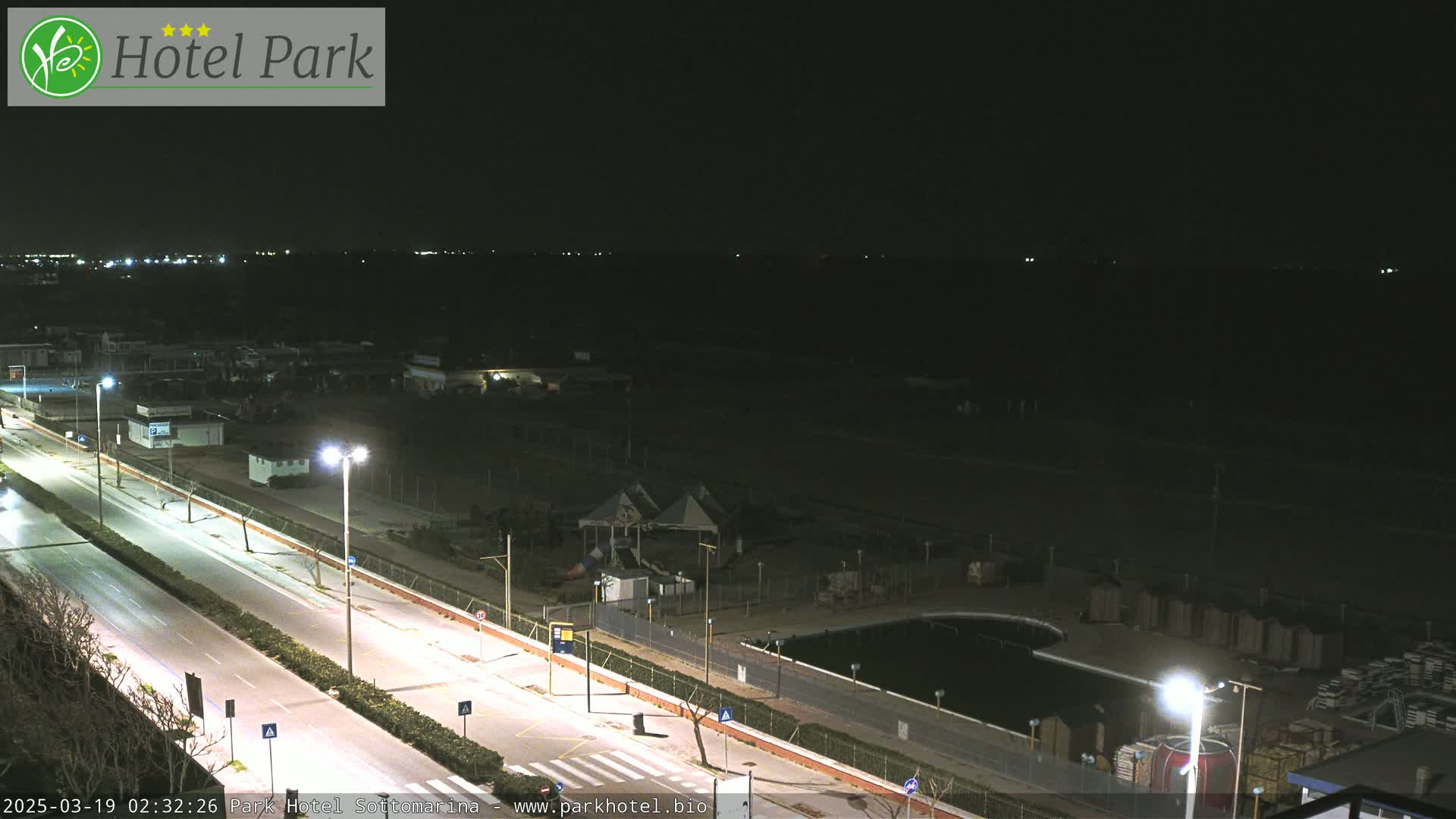 A nighttime, high-angle view of a mostly empty roadway and surrounding area including a small pool and playground, with faint lights visible in the distance.