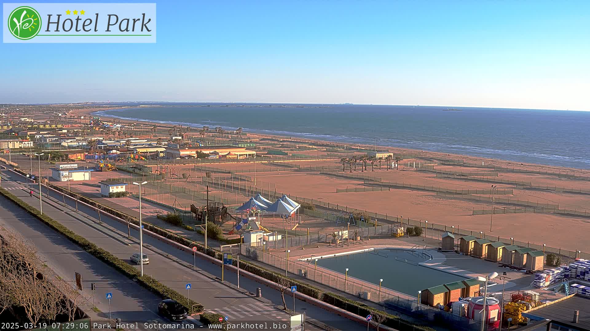 A seaside resort with a swimming pool, playground, and beach under a clear, sunny sky.