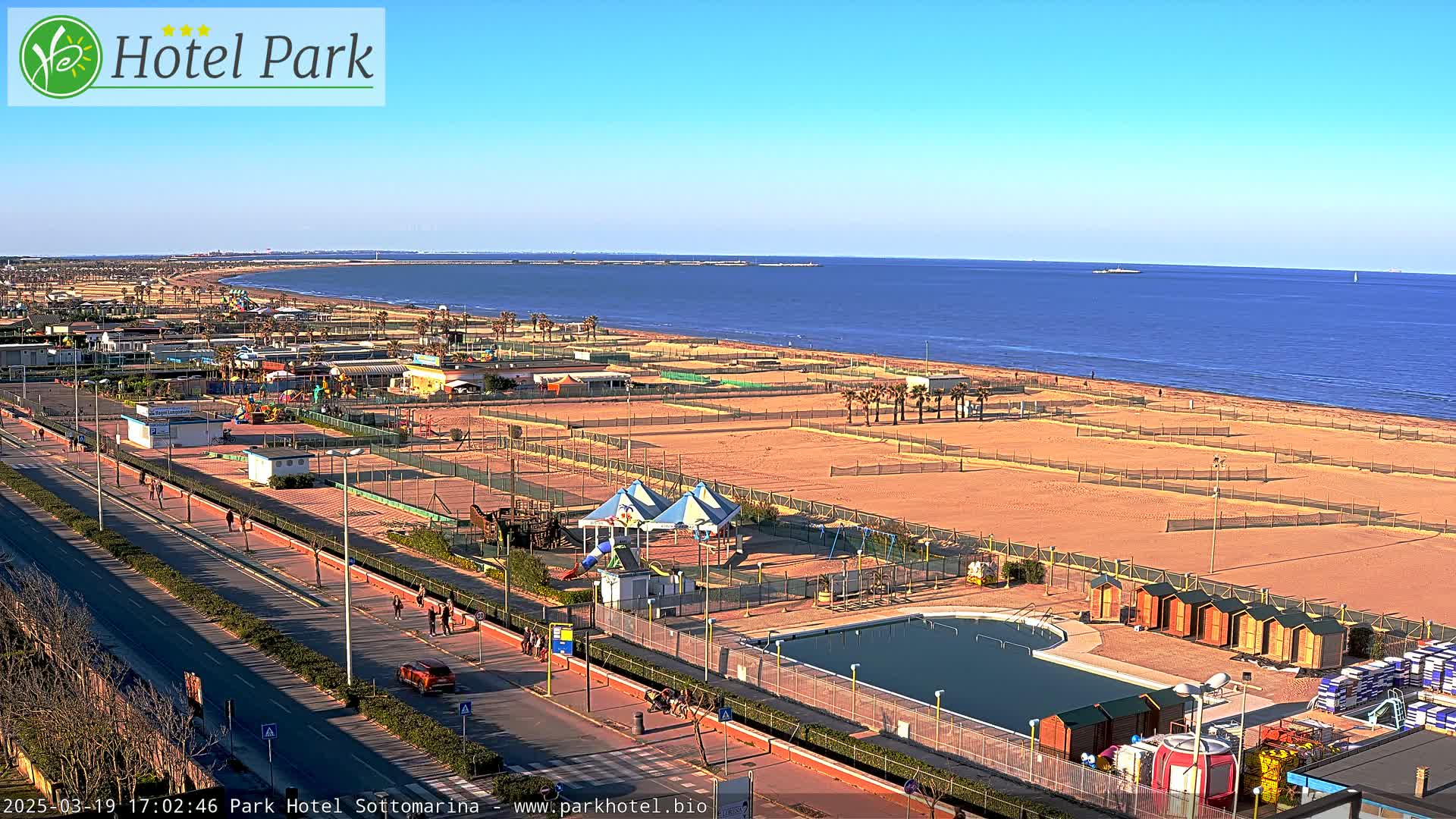 A beachfront view on a sunny day shows a long sandy beach with volleyball courts, a swimming pool, playground equipment, and a road with pedestrians and vehicles.