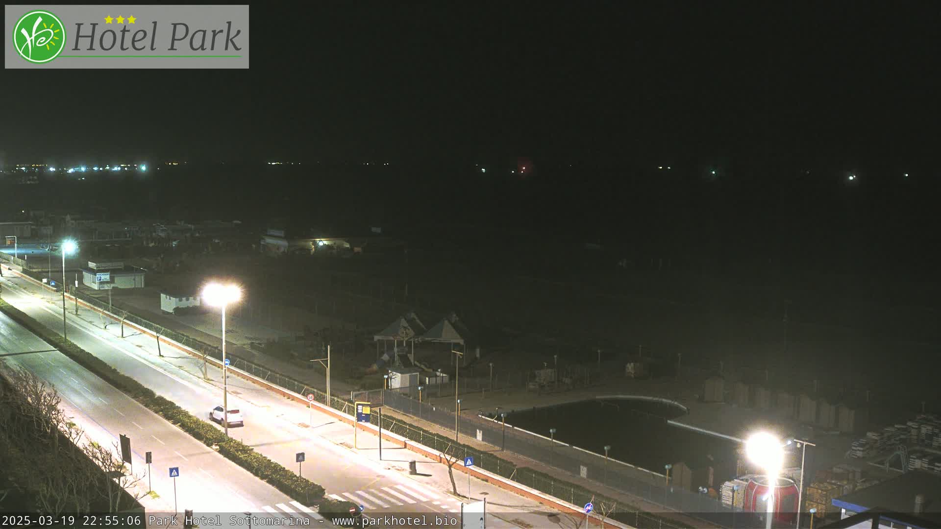 A nighttime view of a coastal road with sparse traffic, adjacent to a mostly dark beachfront area with a few lights and what appears to be a small pool.