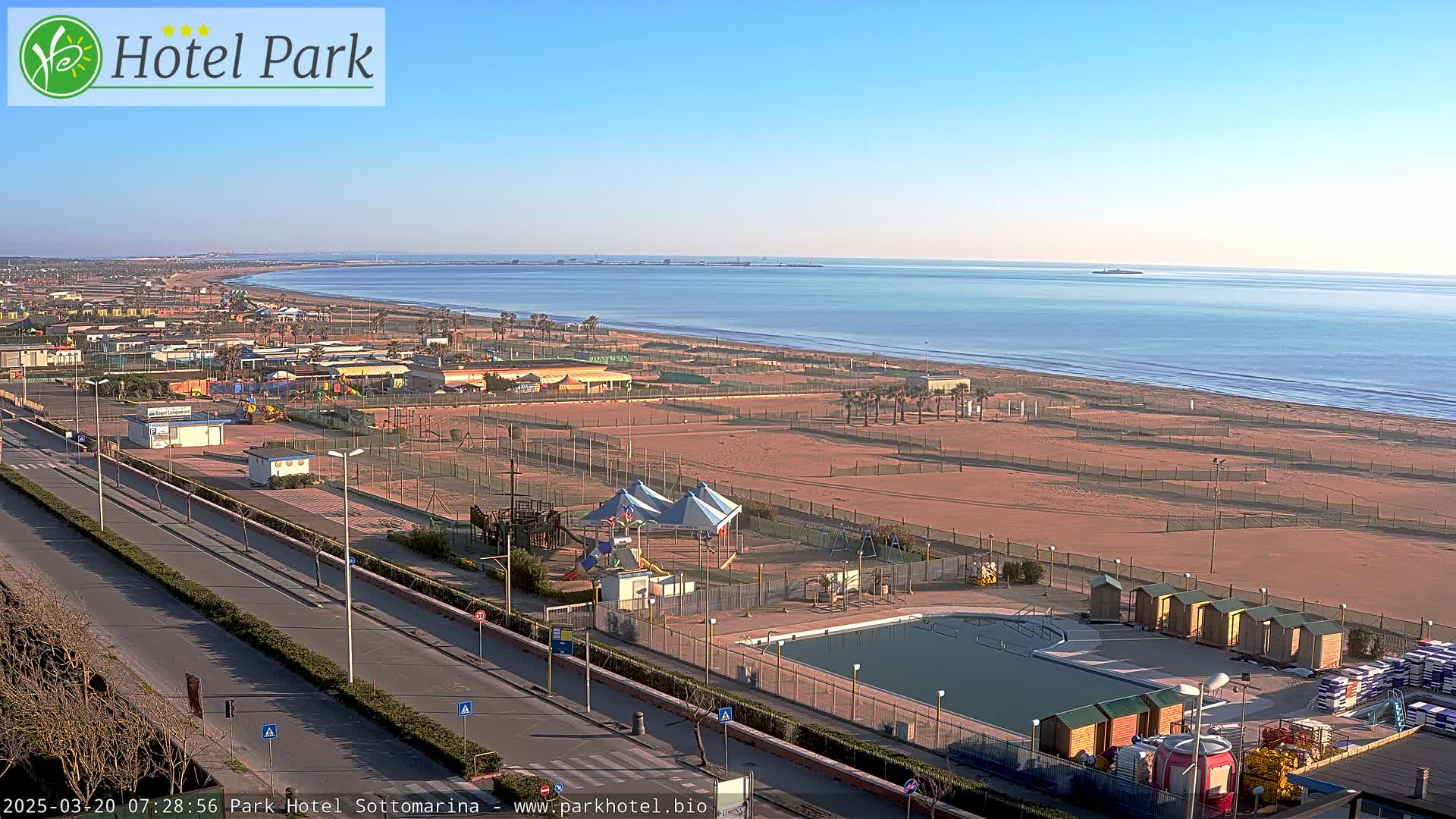 A sunny seaside resort with a swimming pool, playground, and beach, viewed from above.