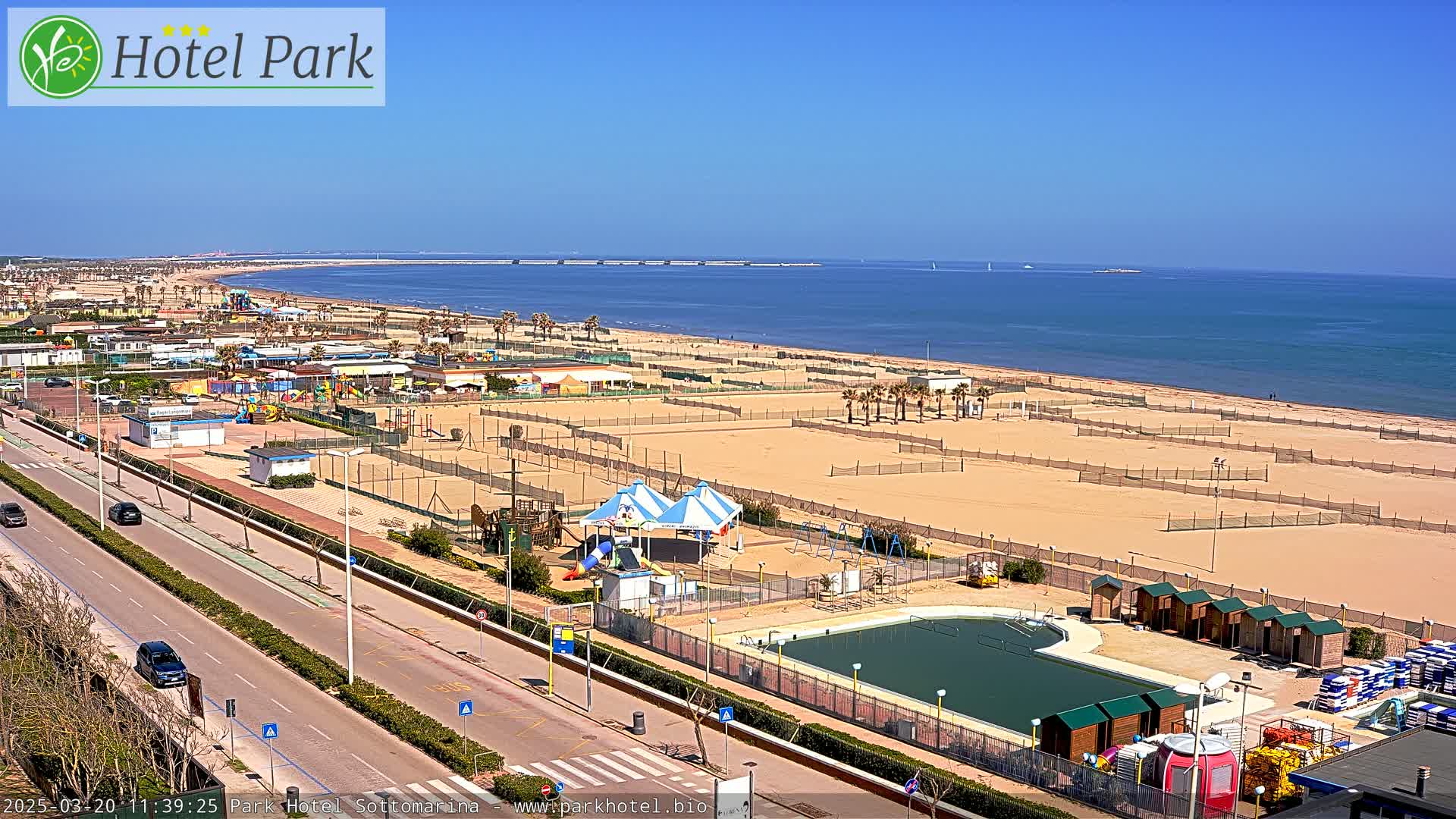 A wide shot on a sunny day shows a beach with volleyball courts, a playground, a pool, and changing rooms, all bordered by a road and palm trees, with a calm ocean in the background.