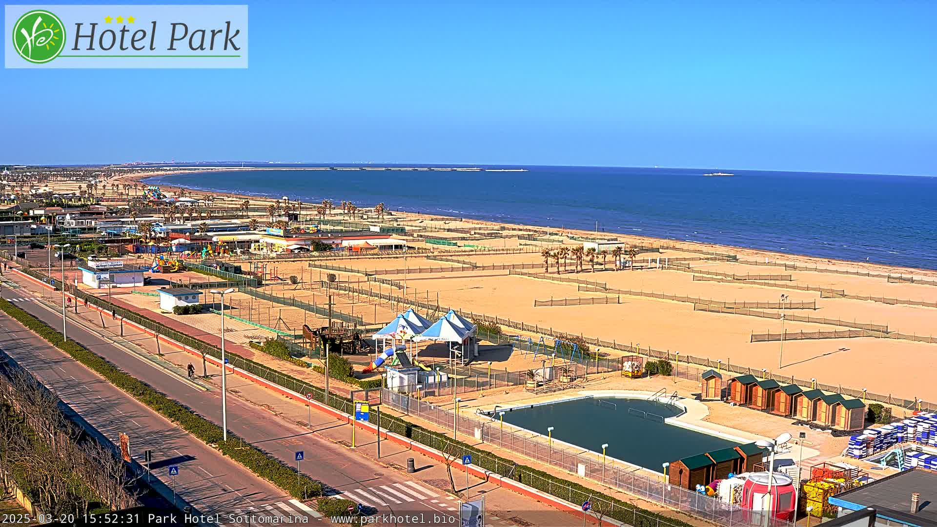 A long sandy beach with volleyball courts, a swimming pool, playground equipment, and beach huts is seen from an elevated viewpoint on a sunny day.