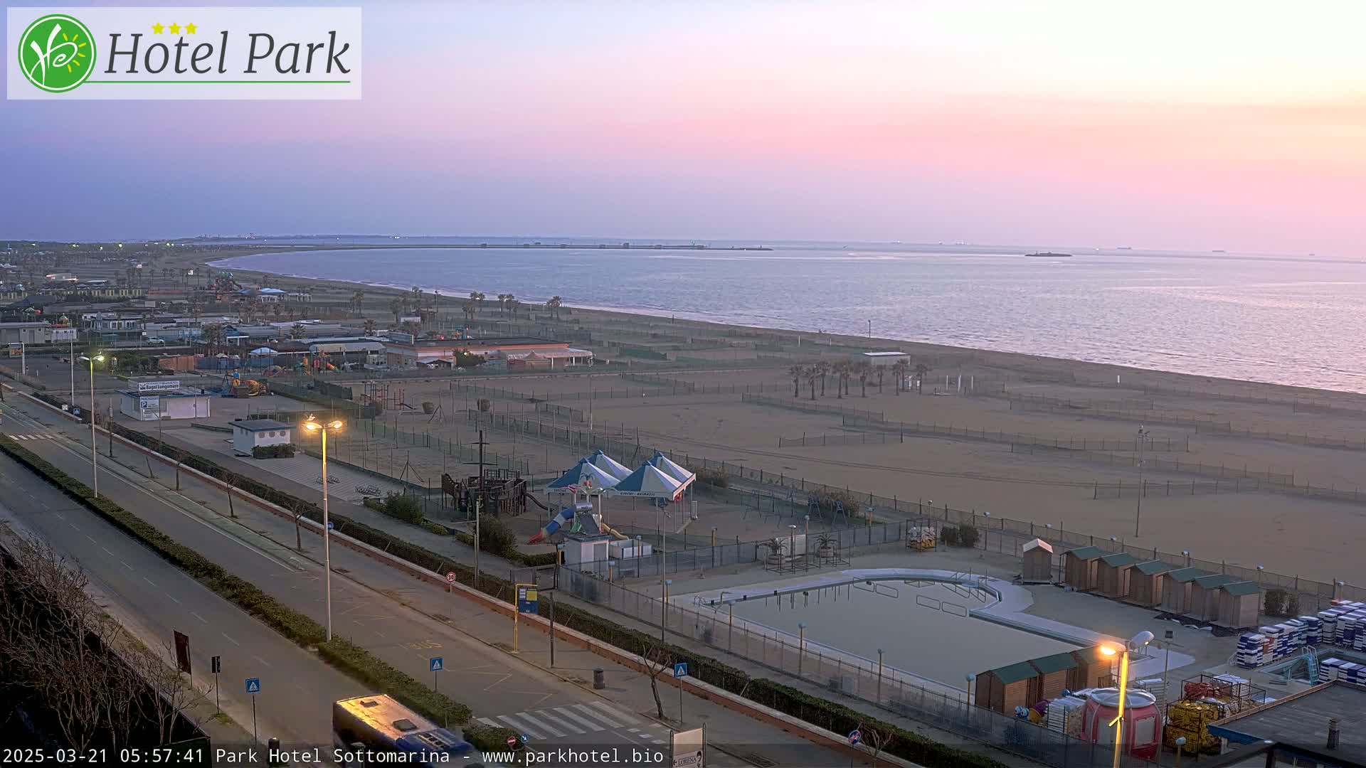 A beach with a playground, swimming pool, and road is seen at dawn under a pink sky.