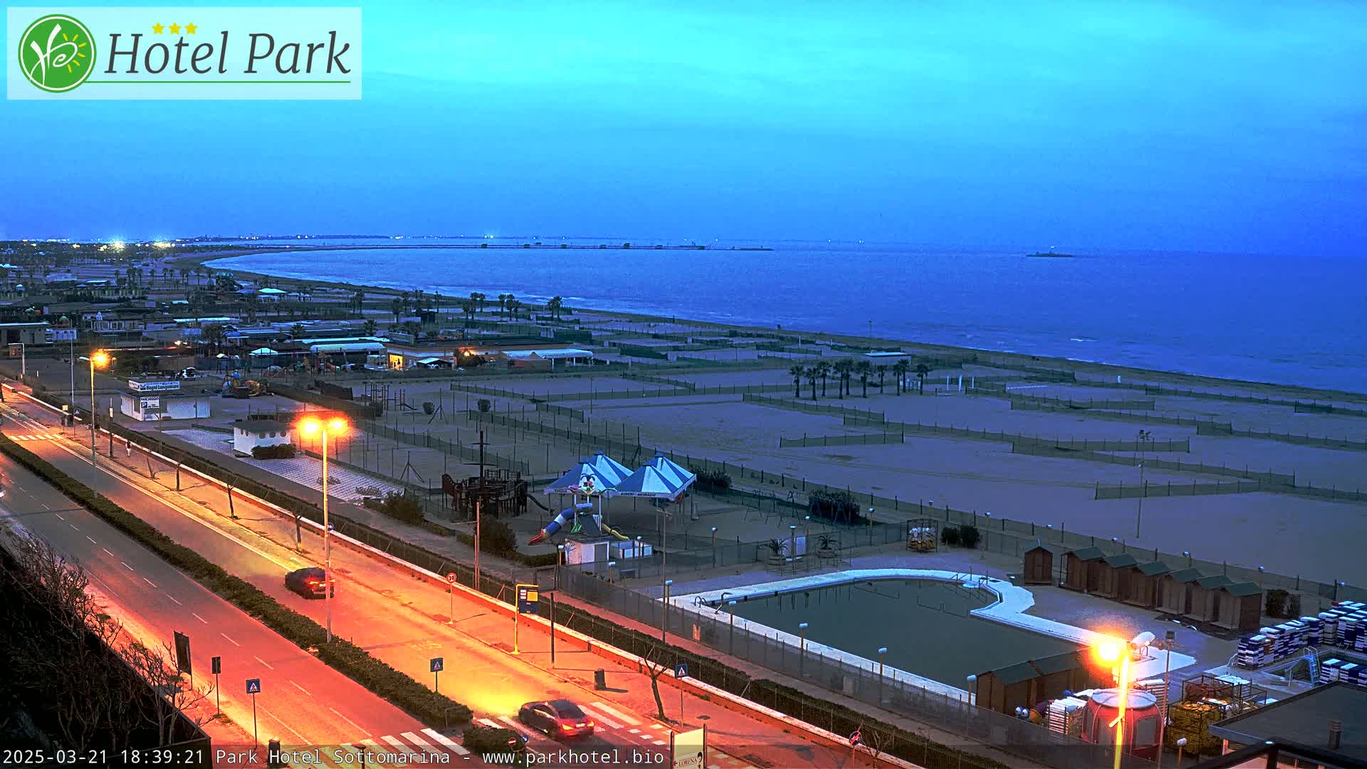 An empty beach at dusk, with a road, playground, and swimming pool visible in the foreground.