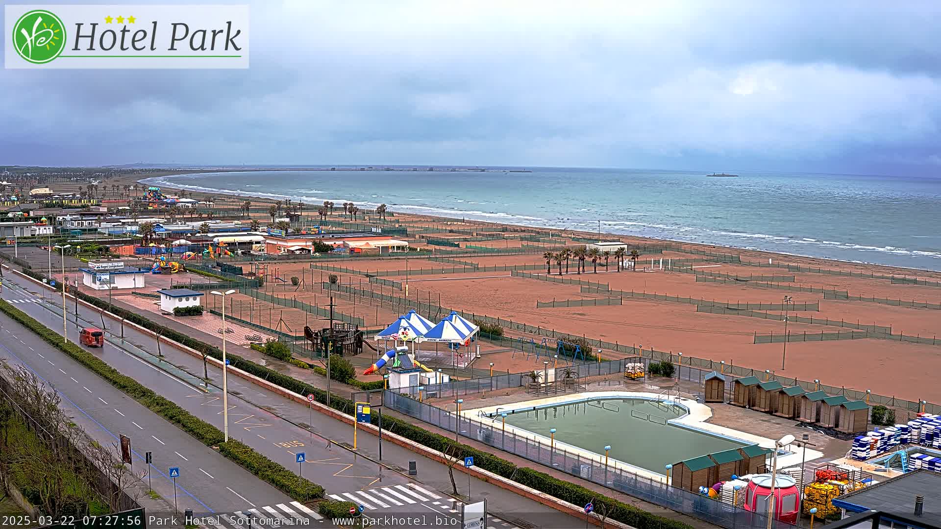 An overcast day reveals a beachfront scene with a swimming pool, playground equipment, and a roadway next to a sandy beach and calm ocean.