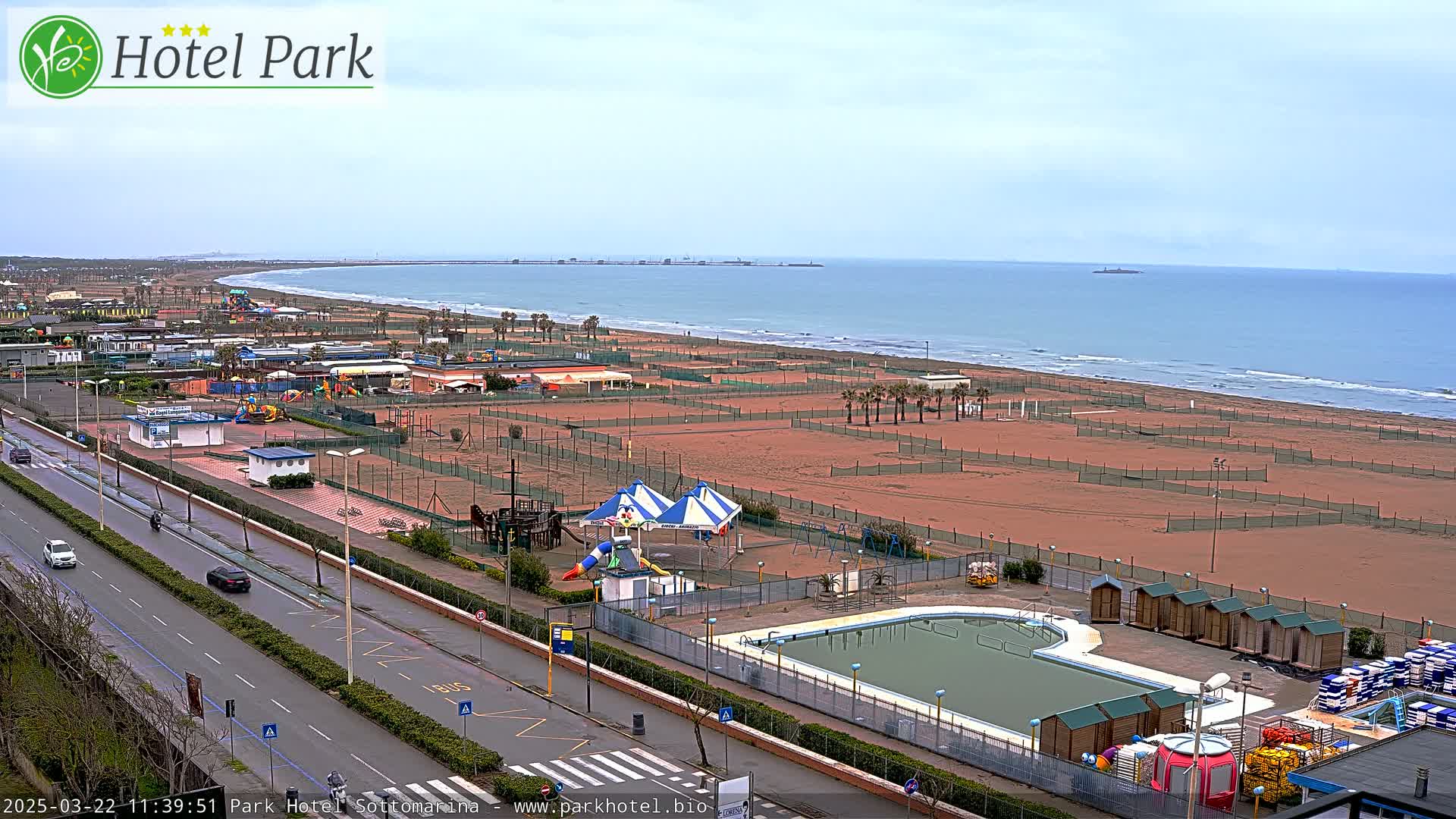 An overcast day reveals a beach with volleyball courts, a swimming pool, playground equipment, and a road running alongside it, all overlooking a calm sea.