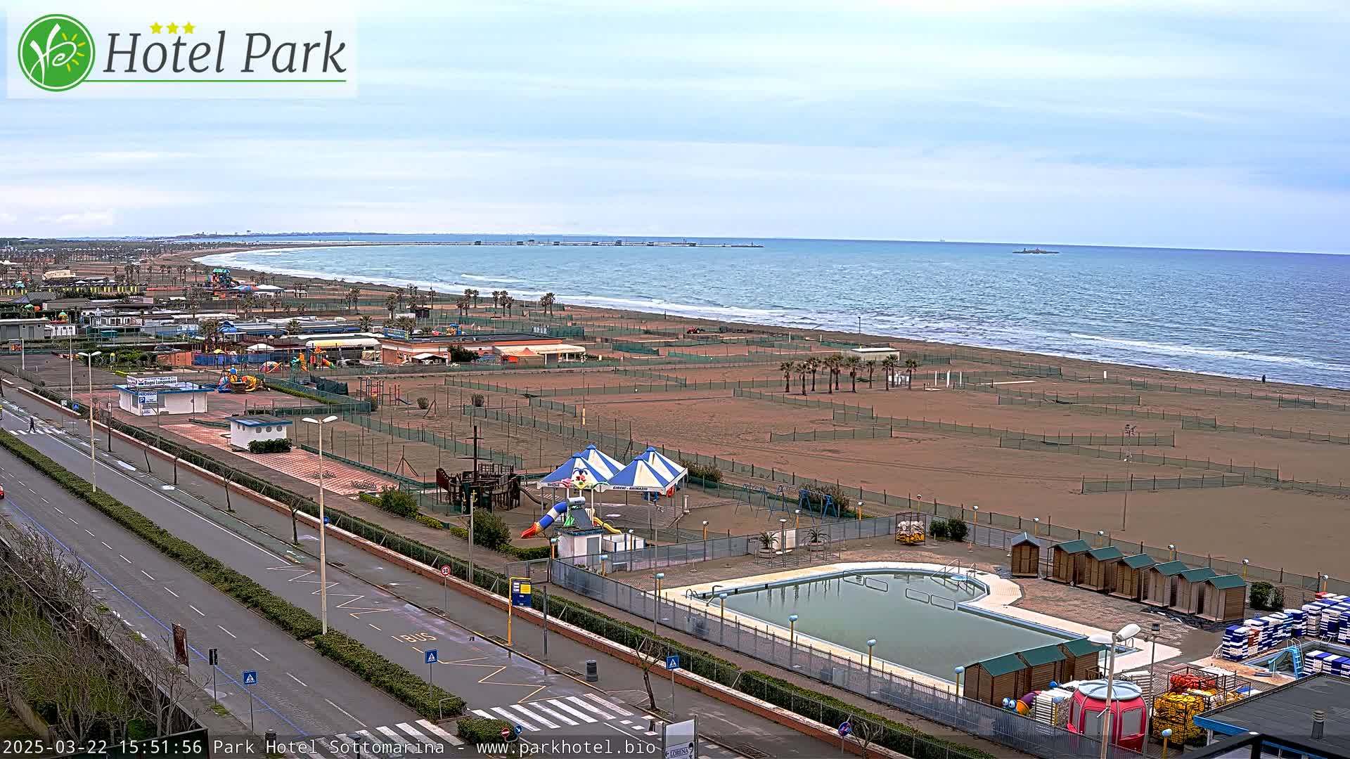 An outdoor view of a beachside area with a swimming pool, playground equipment, and a road, under a partly cloudy sky.
