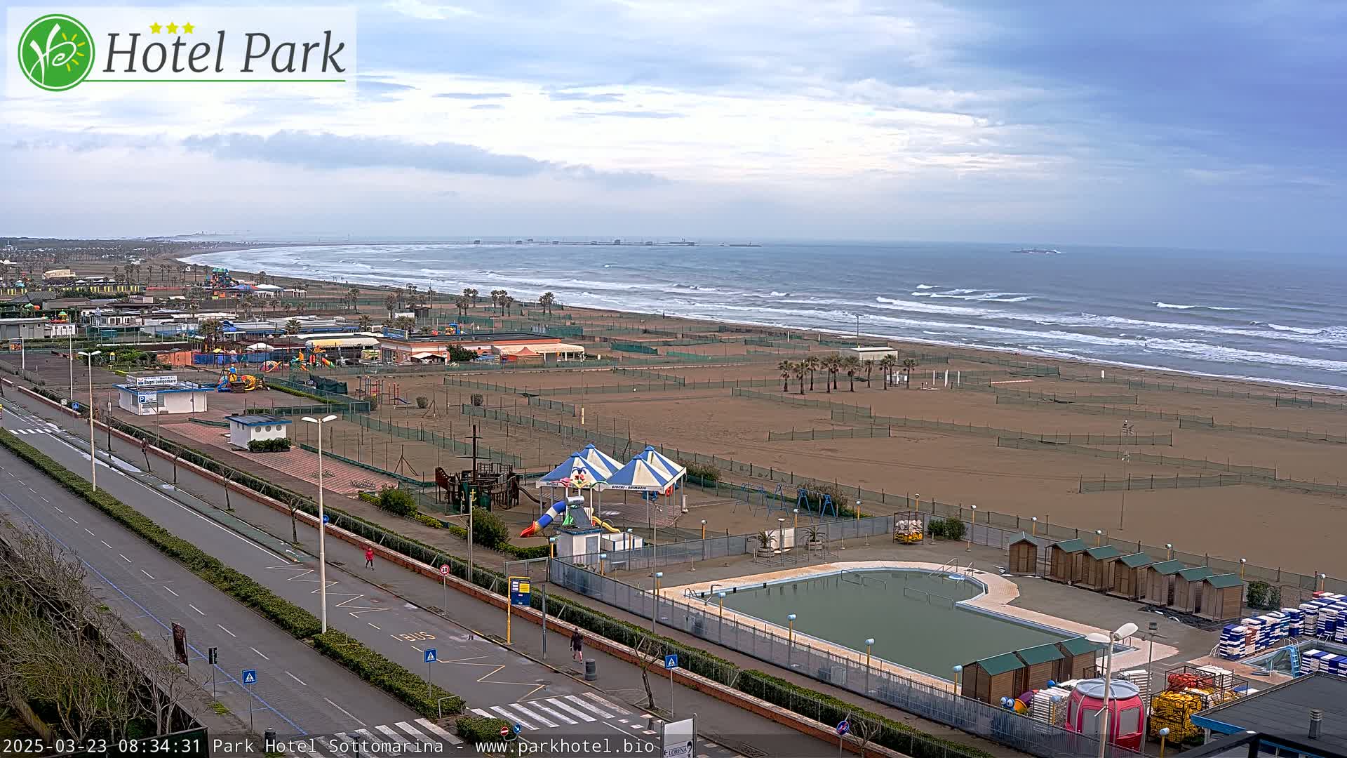 A beachfront area with a swimming pool, playground equipment, and changing rooms, under an overcast sky with waves breaking on the shore.