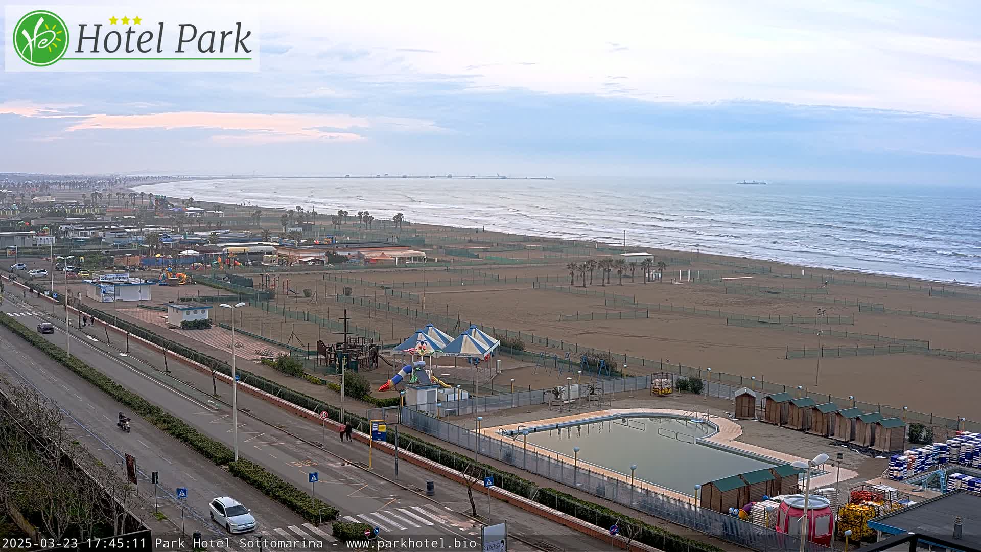 An aerial view of a beach with a swimming pool, playground, and a road, under an overcast sky with a calm sea.