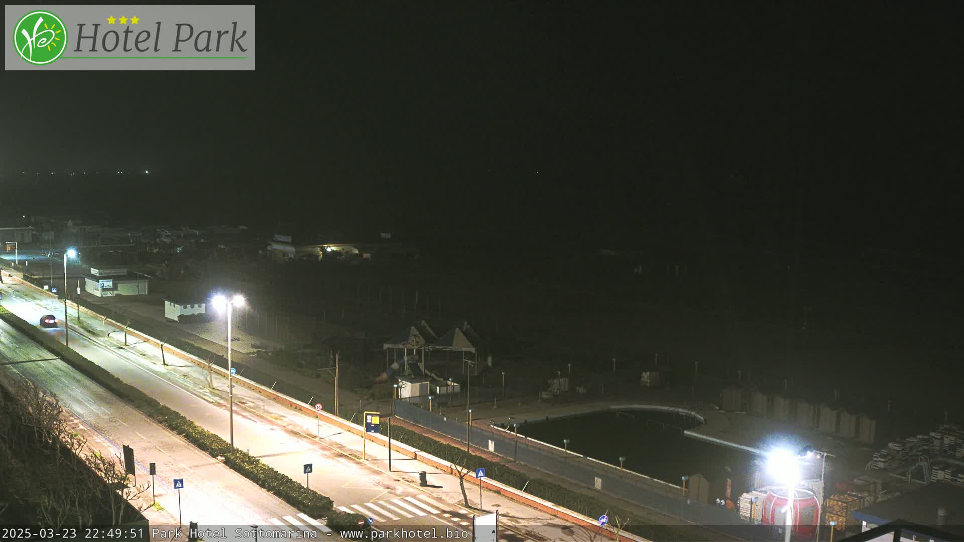 A nighttime view of a mostly empty road and a dark area with a pool and playground, possibly near a beach, under a clear, dark sky.